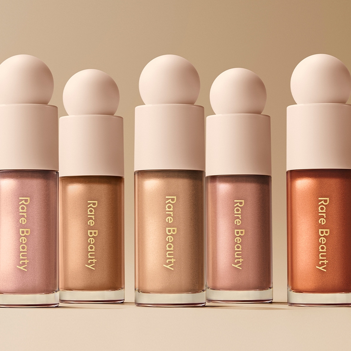 Eine Reihe von Rare Beauty-Highlightern in verschiedenen Farbtönen, von zartem Rosa bis zu warmem Bronze, alle in eleganten, abgerundeten Flaschen.