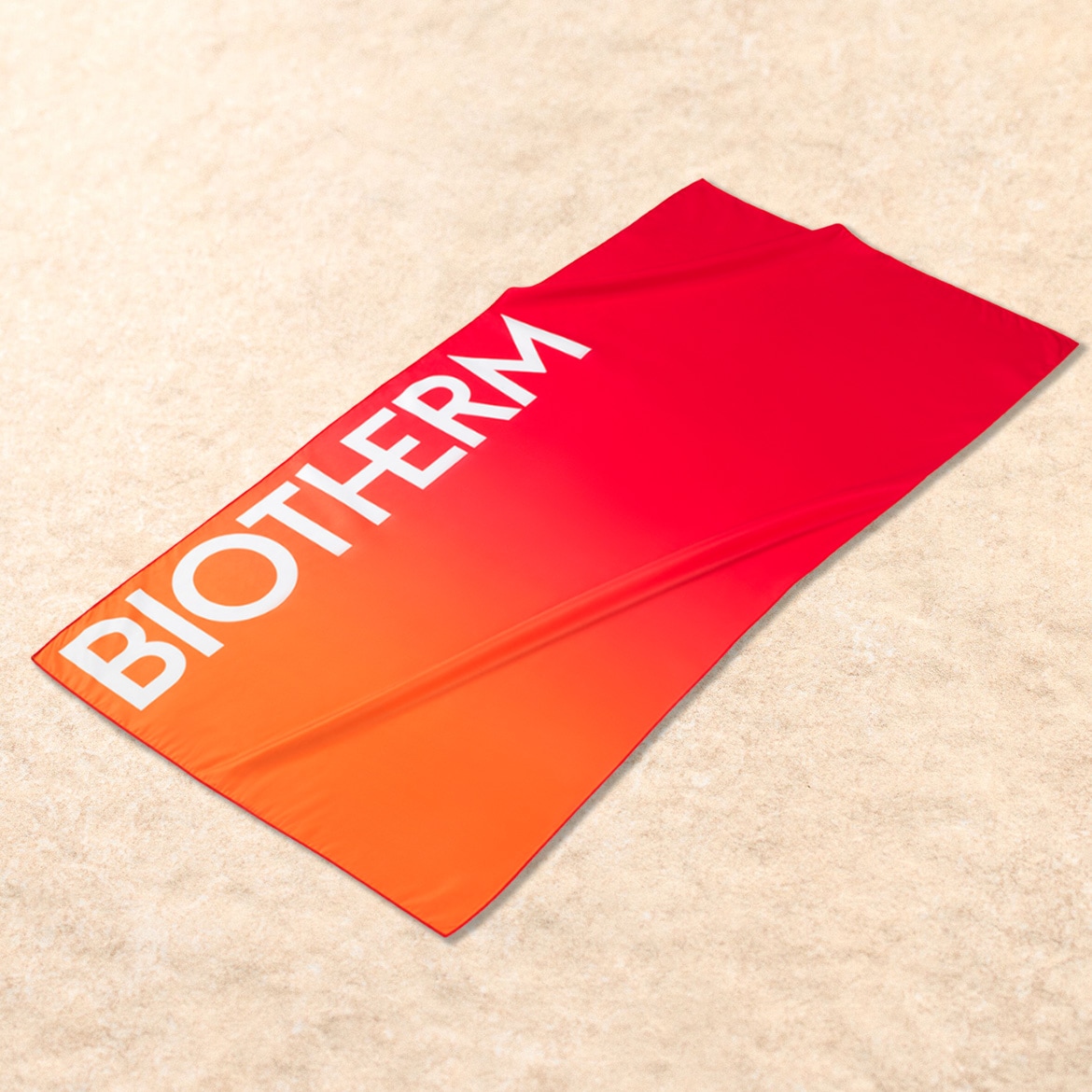 Ein rotes Strandtuch mit orangefarbenem Farbverlauf und dem weißen „BIOTHERM“-Logo liegt auf beigem Sand.
