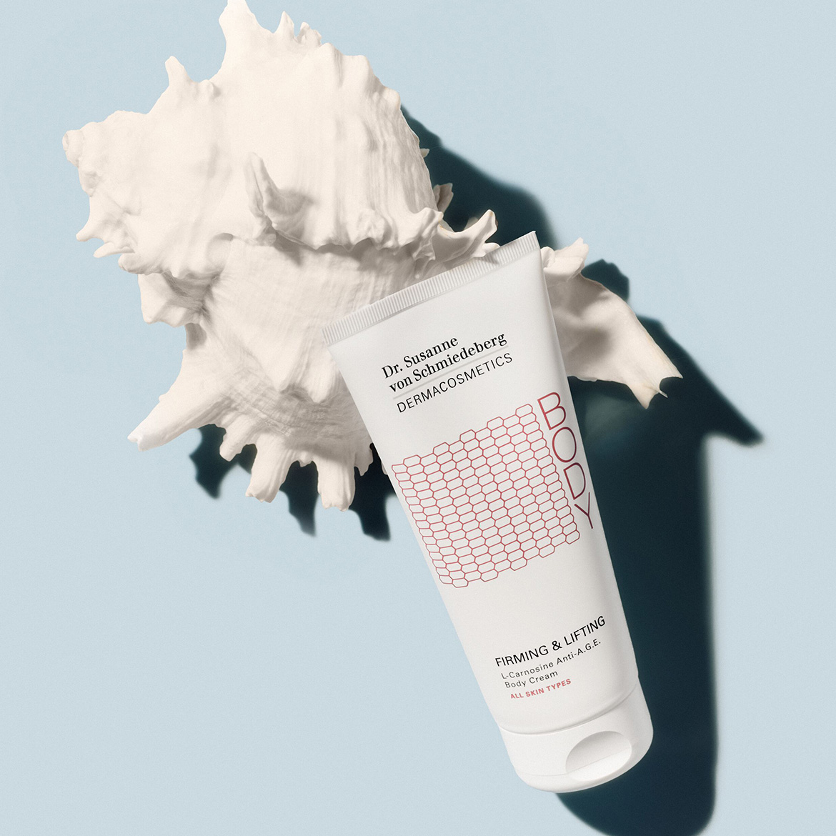 Auf hellblauem Grund liegt eine weiße Tube Dr. Susanne von Schmiedeberg Dermacosmetics Bodylotion mit roter Wabenstruktur und der Aufschrift "Firming & Lifting" neben einer weißen Muschel.