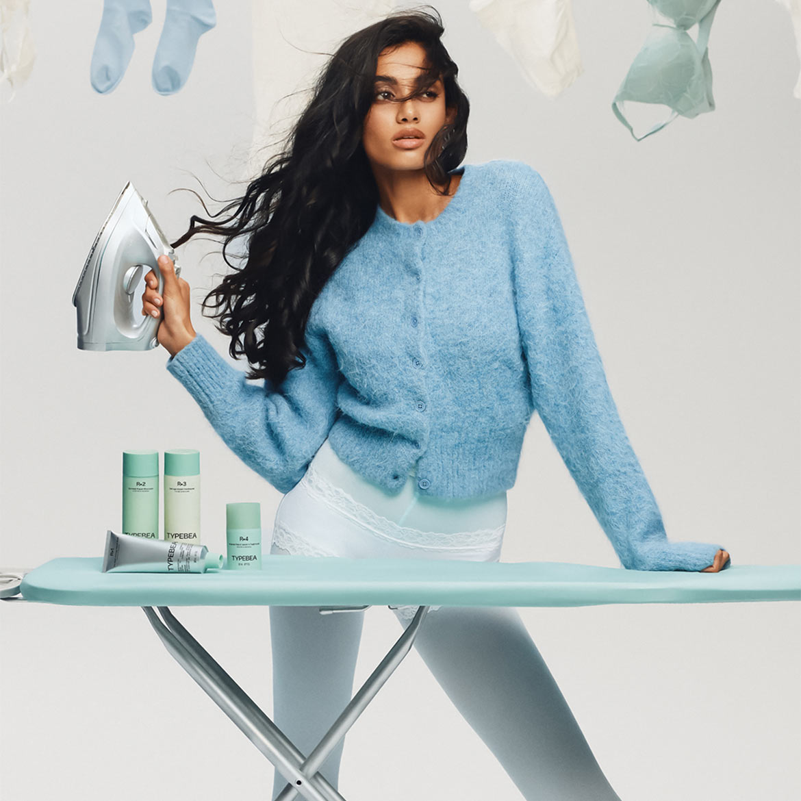 Eine Frau mit langen, dunklen Haaren, die einen blauen Cardigan trägt, hält ein Bügeleisen in der Hand, während sie an einem Bügelbrett mit Hautpflegeprodukten der Marke TYPEBEA steht.