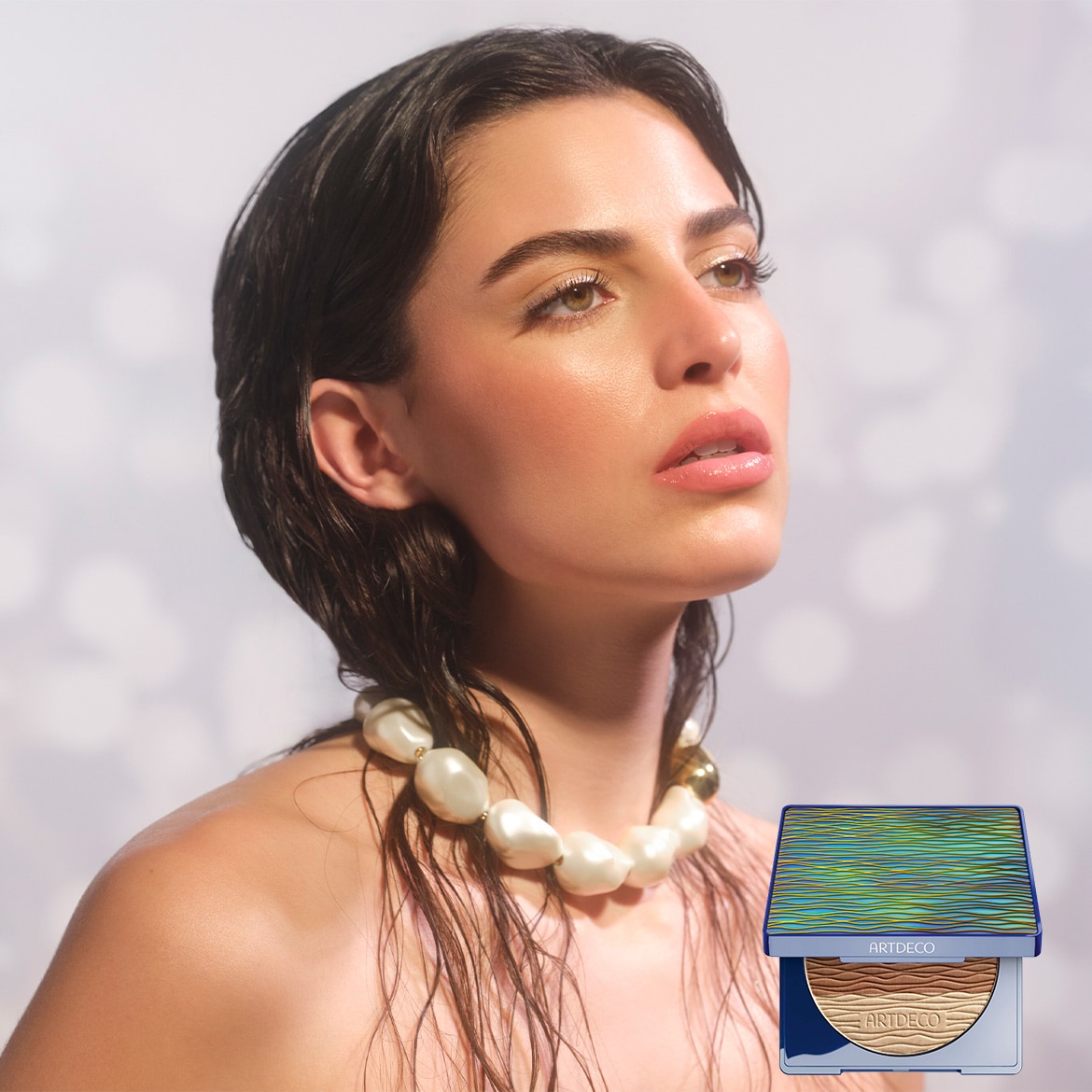 Nahaufnahme einer Frau mit nassen, dunklen Haaren und einer Perlenkette, die ein Artdeco-Produkt präsentiert, das in einer blaugrünen Dose mit gewelltem Muster und einem bronzefarbenen Puder enthalten ist.