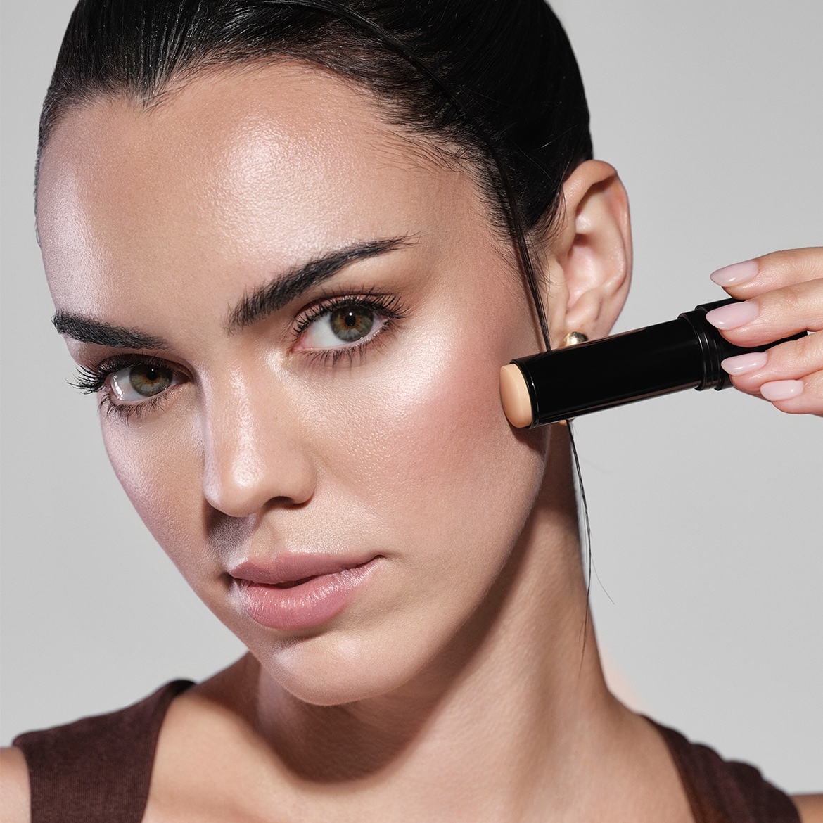 Eine Frau mit olivfarbenen Augen und zurückgekämmtem Haar trägt einen beigen Concealer-Stift auf ihre Wange auf.