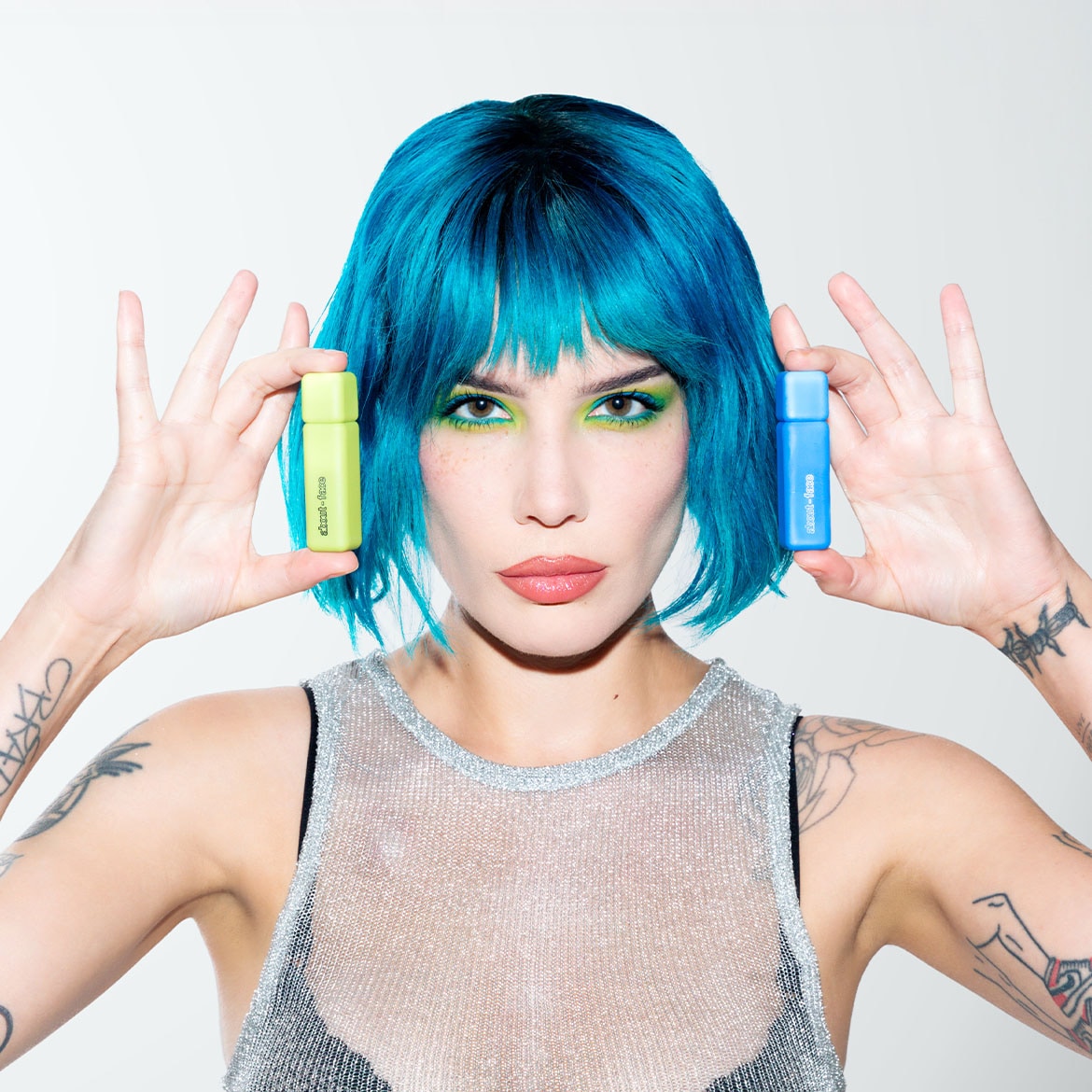 Eine Frau mit blauen Haaren und grünem Lidschatten hält eine hellgrüne und eine blaue Flasche mit der Aufschrift „about-face“ in den Händen.