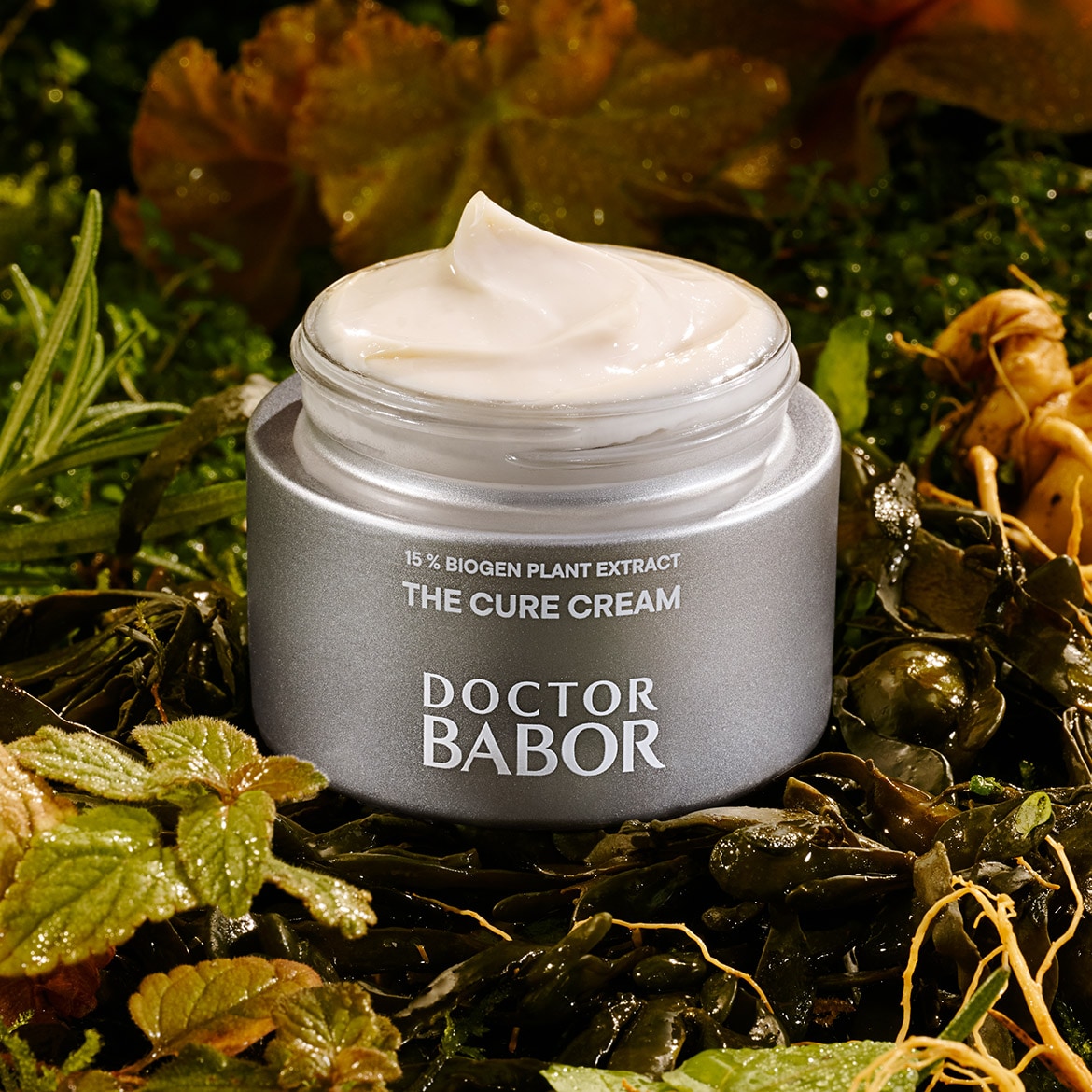 Die Doctor Babor The Cure Cream mit 15% Biogen Pflanzenextrakt in einem silbernen Tiegel mit klarer Oberseite, umgeben von üppigem Grün.