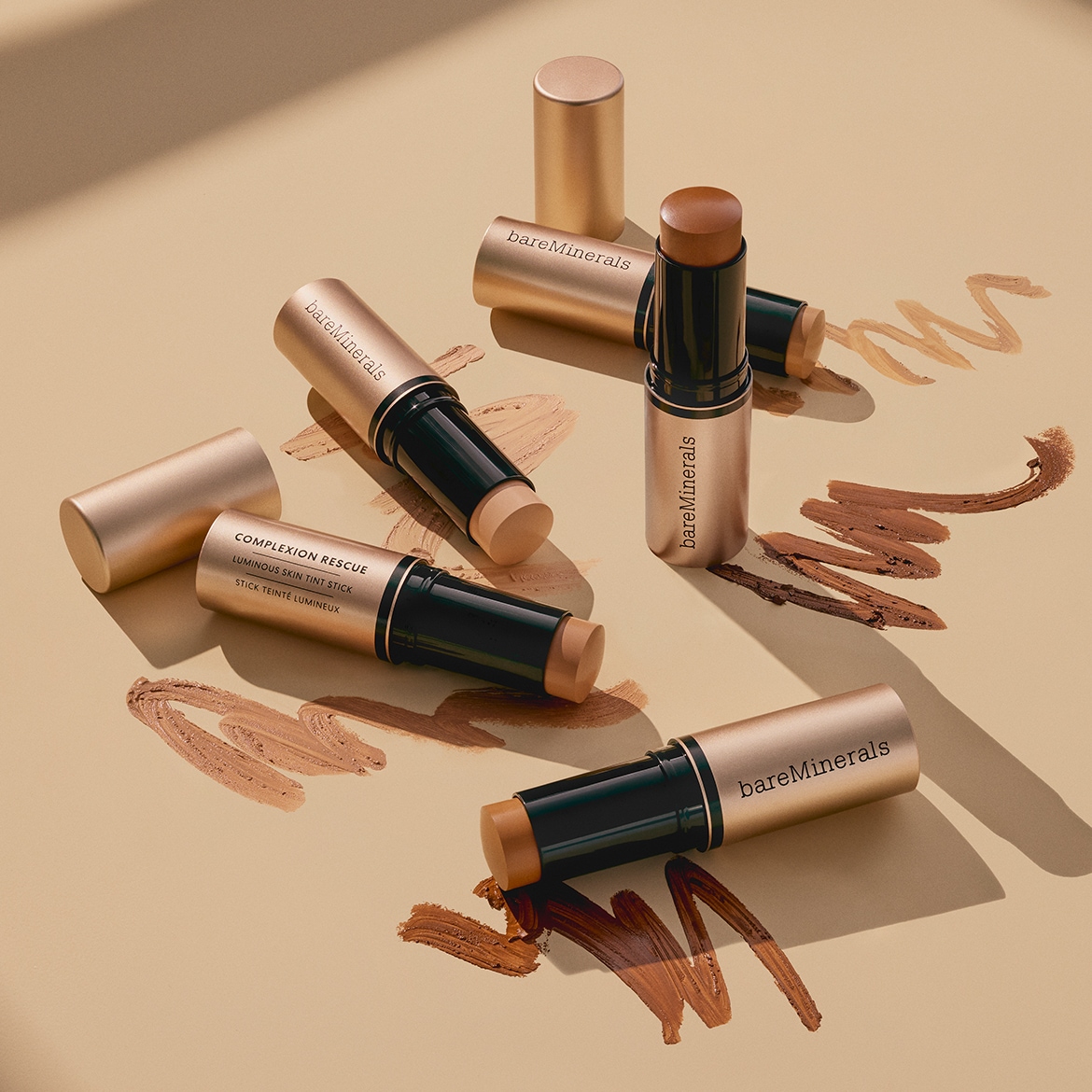 Entdecken Sie die bareMinerals Complexion Rescue Luminous Skin Tint Sticks in verschiedenen Farbtönen, die auf einem beigefarbenen Hintergrund mit Farbwischern präsentiert werden.
