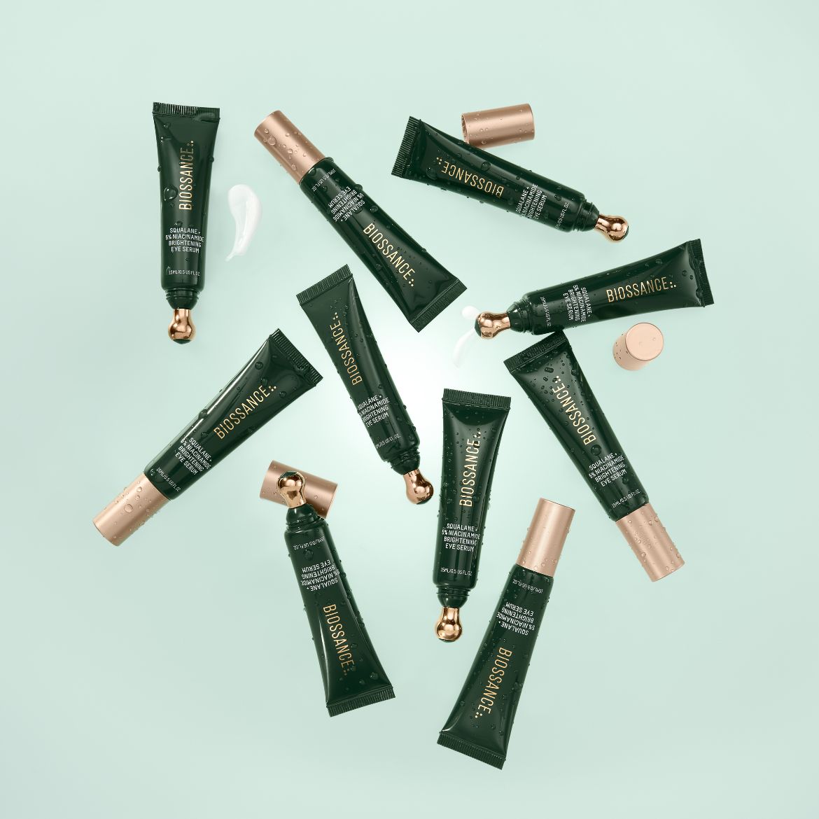 Mehrere Tuben des Biossance Squalane + 5% Niacinamide Brightening Eye Serum sind auf einem hellgrünen Hintergrund verstreut, einige mit goldenen Applikatoren und sichtbaren Cremespuren.