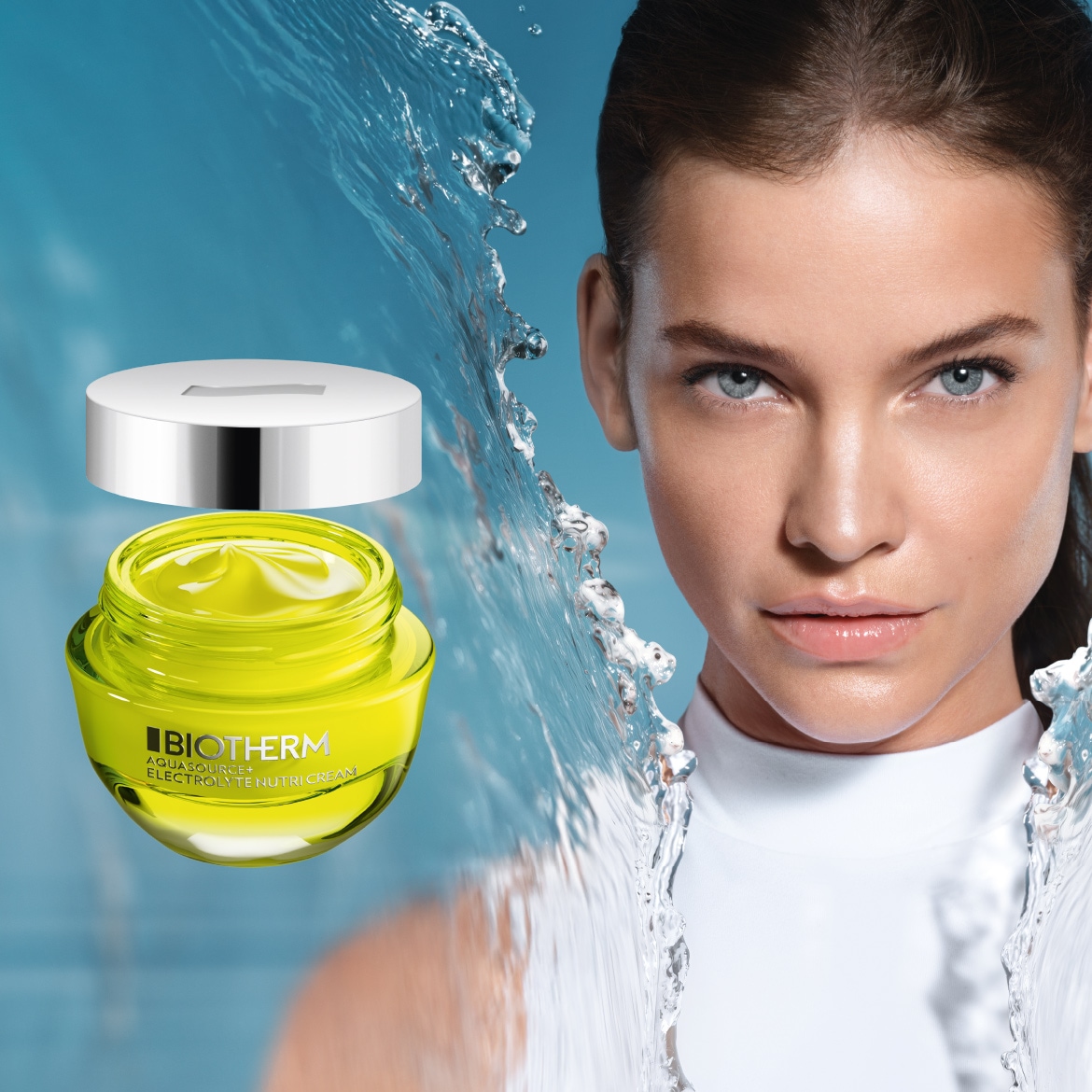 Eine Frau mit makelloser Haut blickt direkt in die Kamera, während ein offener Tiegel Biotherm Aquasource Elektrolyt Nutri Cream mit leuchtend gelber Creme und silbernem Deckel vor einem Hintergrund aus spritzendem Wasser zu sehen ist.