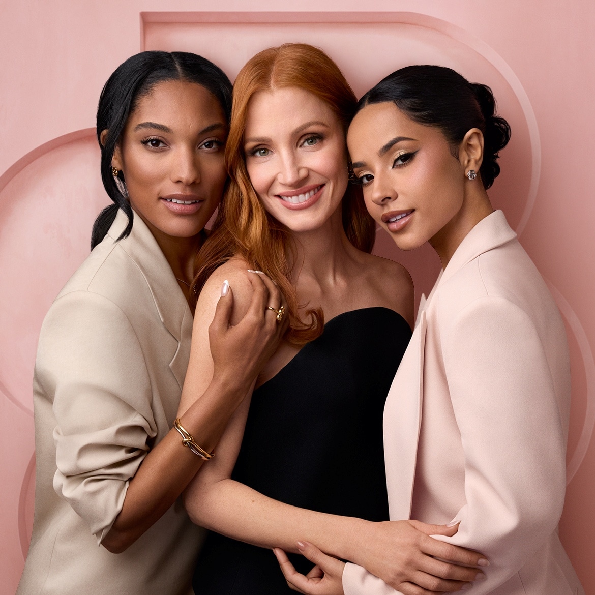 Drei Frauen, darunter Jessica Chastain, posieren lächelnd vor einem rosafarbenen Hintergrund, wobei die Frau in der Mitte ein schwarzes, trägerloses Oberteil trägt und die anderen beiden helle Blazer.
