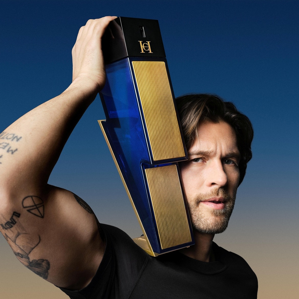 Ein Mann mit braunen Haaren hält eine blaue und goldene Carolina Herrera Bad Boy Duftflasche in Blitzform über seiner Schulter.