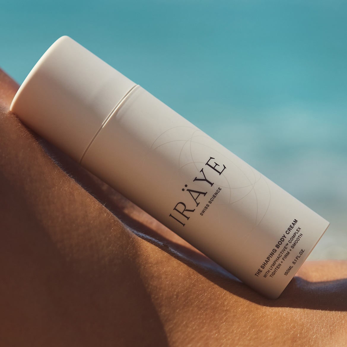 A white tube of IRAYE Swiss Science The Shaping Body Cream with Lymphactive Complex, promising to tighten, firm, and smooth skin, against a backdrop of skin and blue sky.
