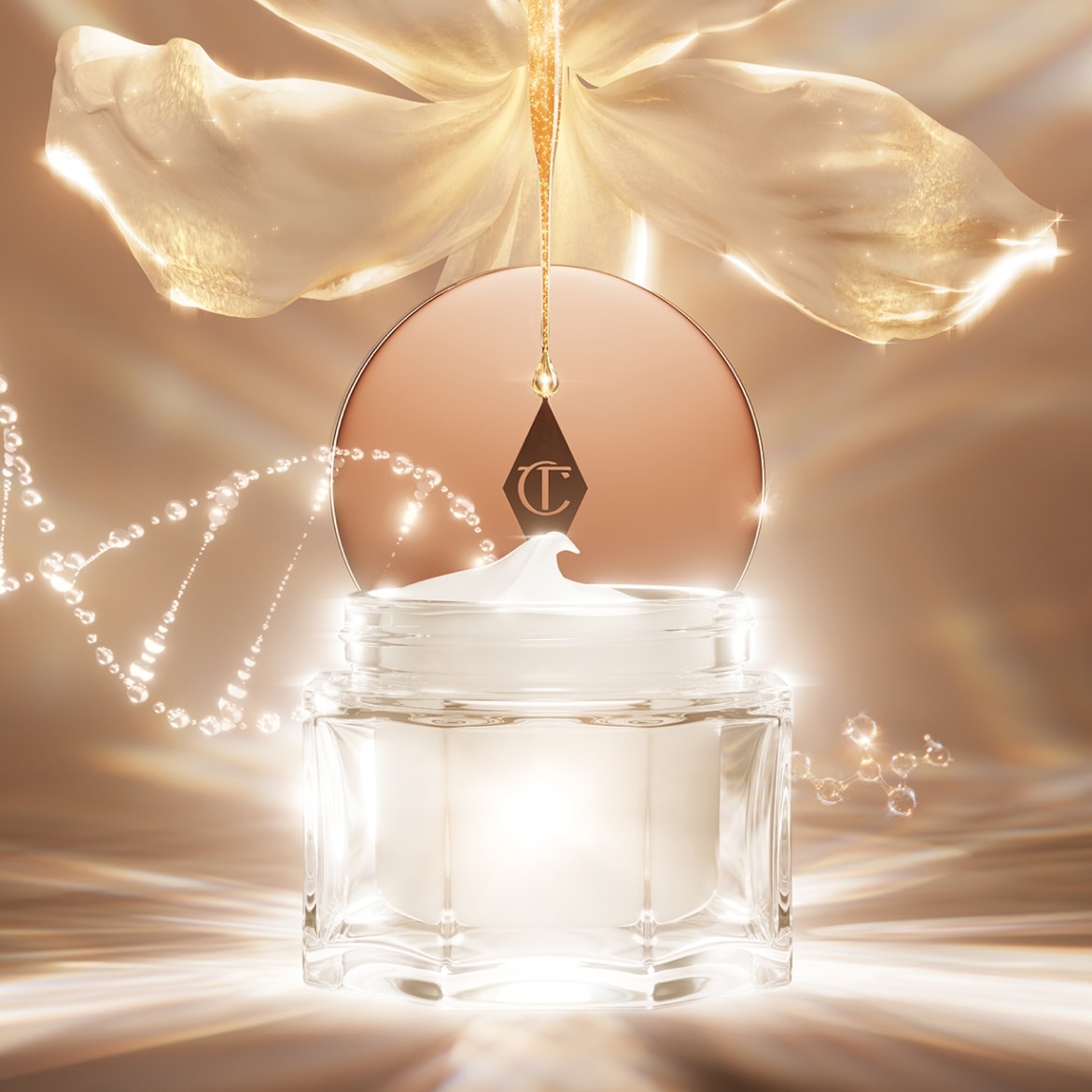 Nahaufnahme einer durchsichtigen Glasdose mit weißer Creme, die mit einem roségoldenen Deckel mit dem Charlotte Tilbury-Logo verschlossen ist, mit einer goldenen Blume und einer goldenen Flüssigkeit im Hintergrund.