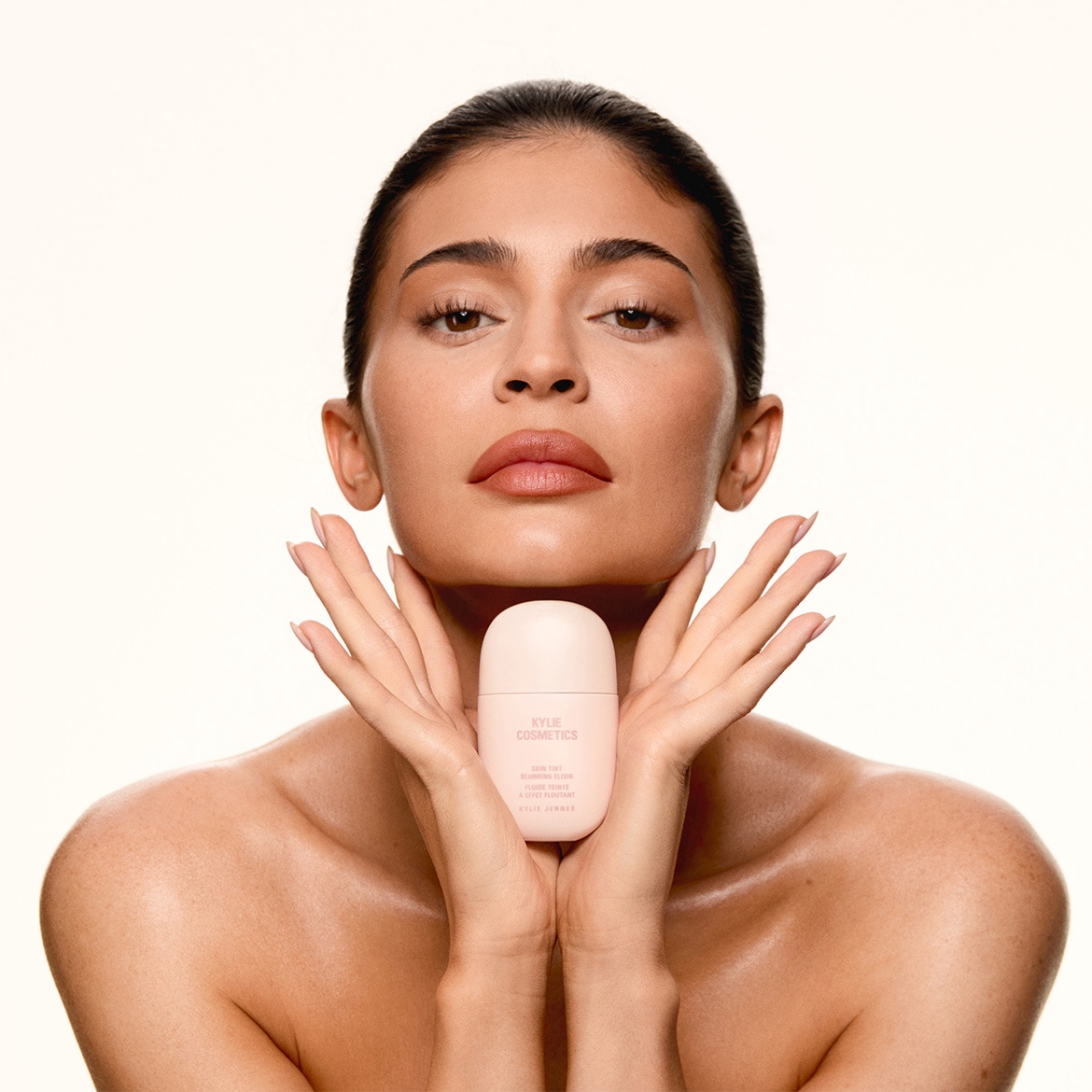 Die Frau hält eine hellrosa Kylie Cosmetics Skin Tint Flasche in ihren Händen, die eine getönte Flüssigkeit für einen strahlenden Effekt verspricht.