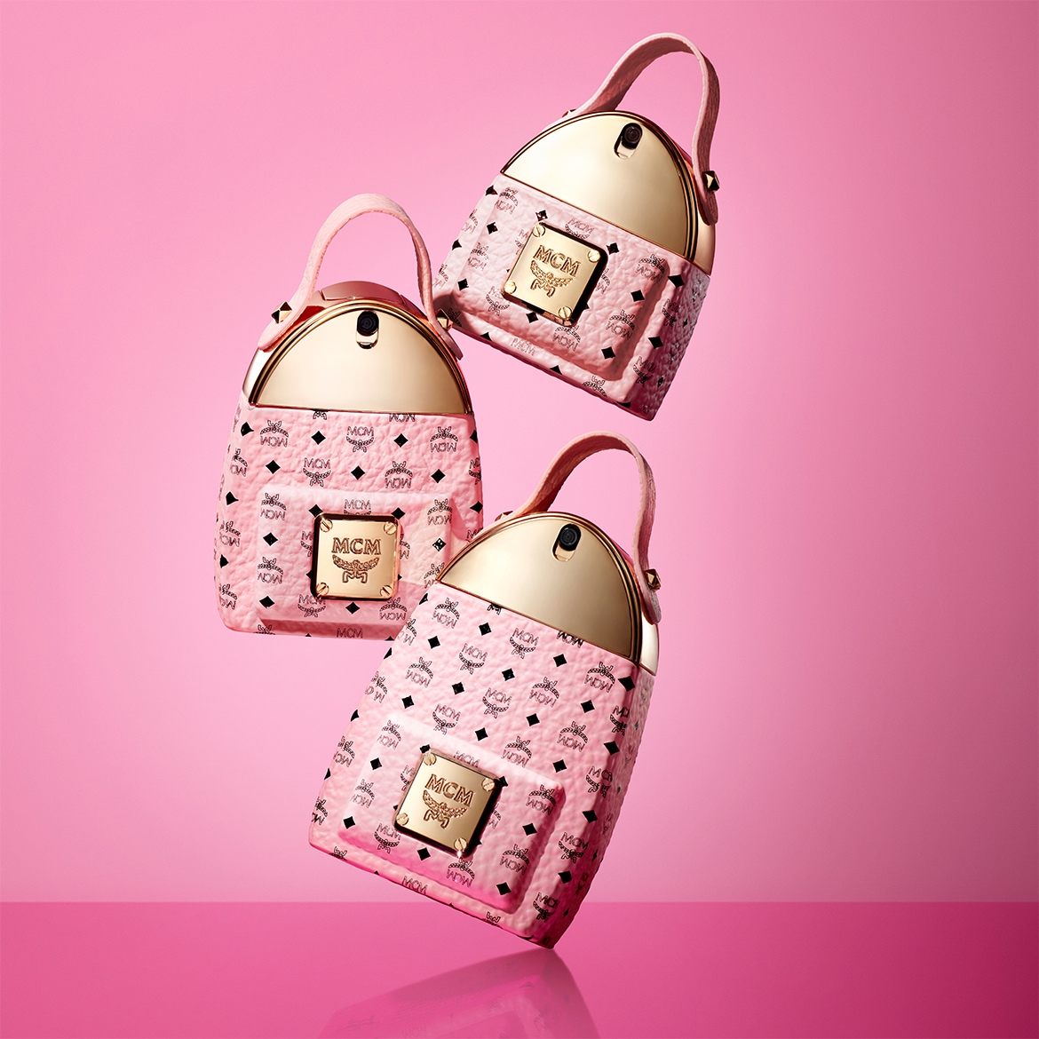 Drei MCM-Handtaschen in Rosa mit goldenen Akzenten schweben vor einem pinkfarbenen Hintergrund und zeigen das ikonische MCM-Logo.