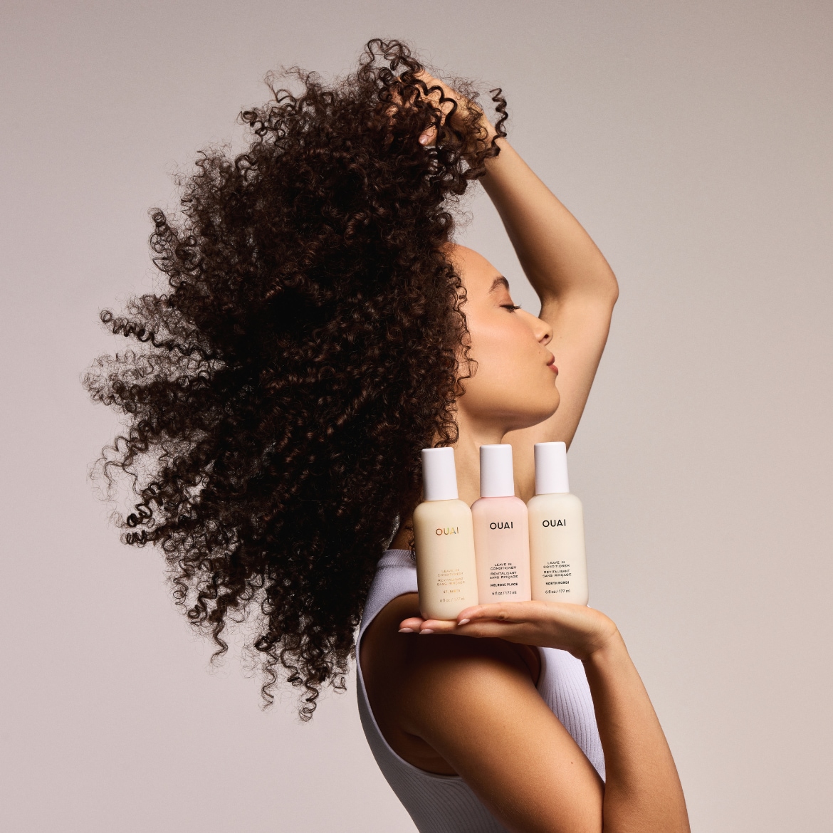 Eine Frau mit lockigem Haar hält drei OUAI Leave-In Conditioner-Sprays in den Farben Beige, Rosa und Hellbeige in der Hand.