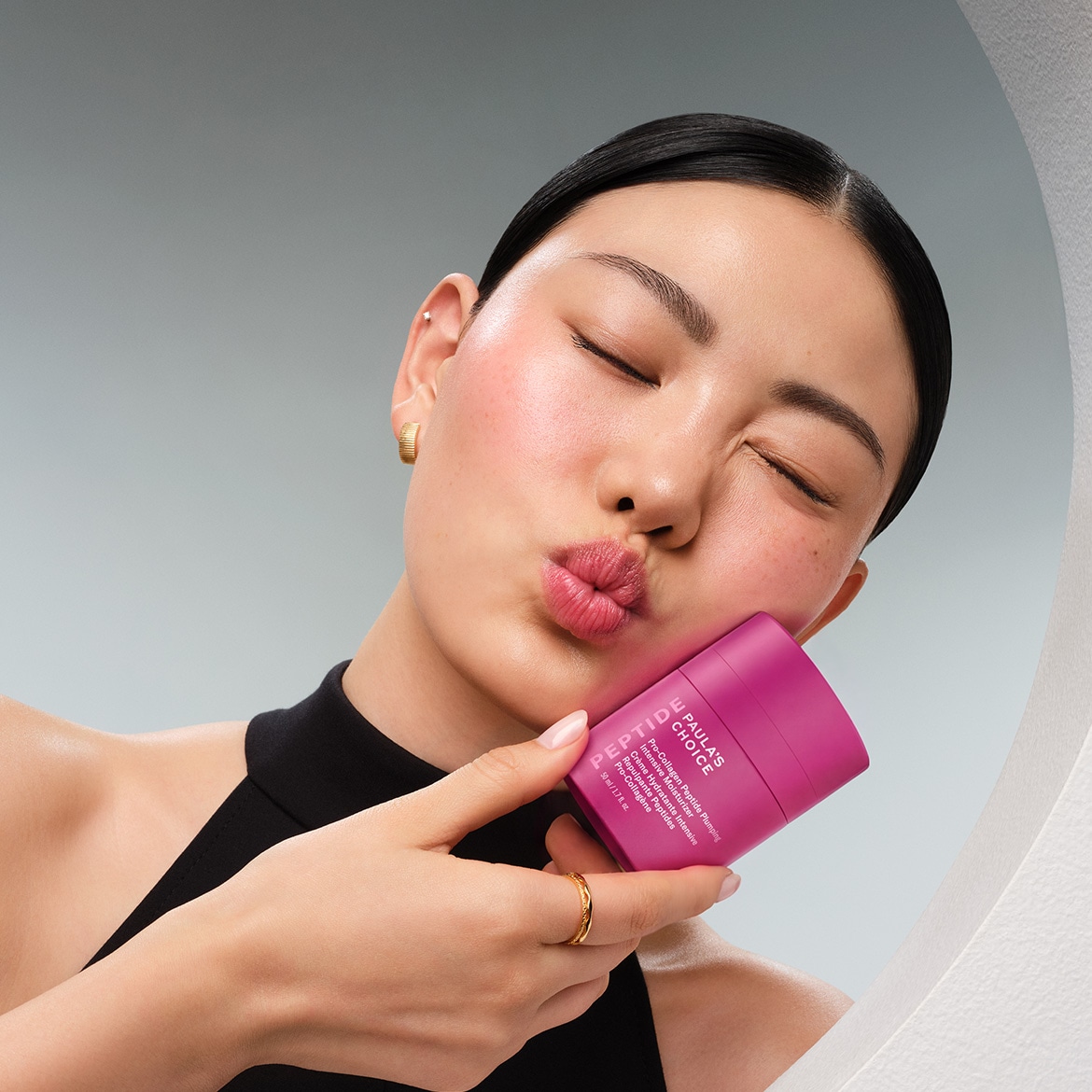Eine Frau mit geschlossenen Augen und einem Kussmund hält eine Paula's Choice Pro-Collagen Peptide Plumping Intensive Moisturizer in einem leuchtenden Pinkton, die 50 ml des Produkts enthält.