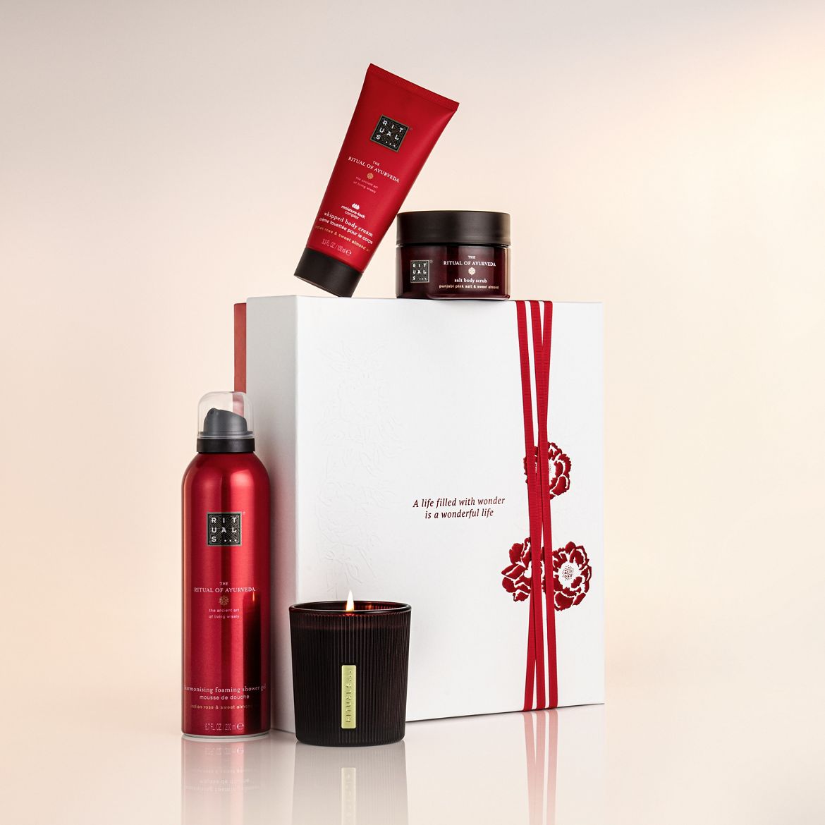 The Ritual of Ayurveda set includes body cream, scrub, shower gel, and candle, presented in red packaging and a white gift box with red ribbon and floral design.