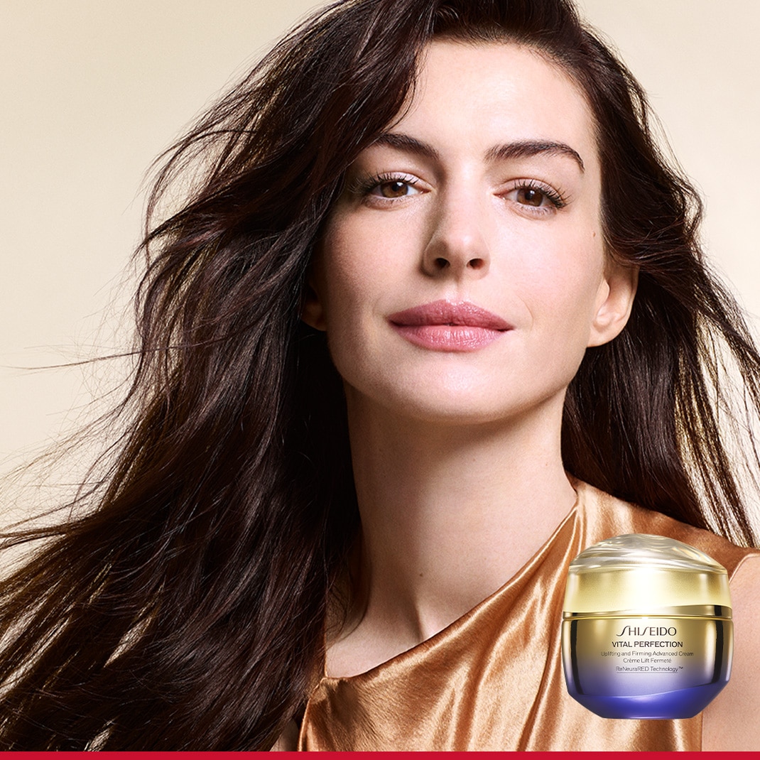 Strahlende Frau mit dunklem, gewelltem Haar und einem goldenen Oberteil, neben der die Shiseido Vital Perfection Lifting Advanced Creme in einem golden-violetten Tiegel steht.