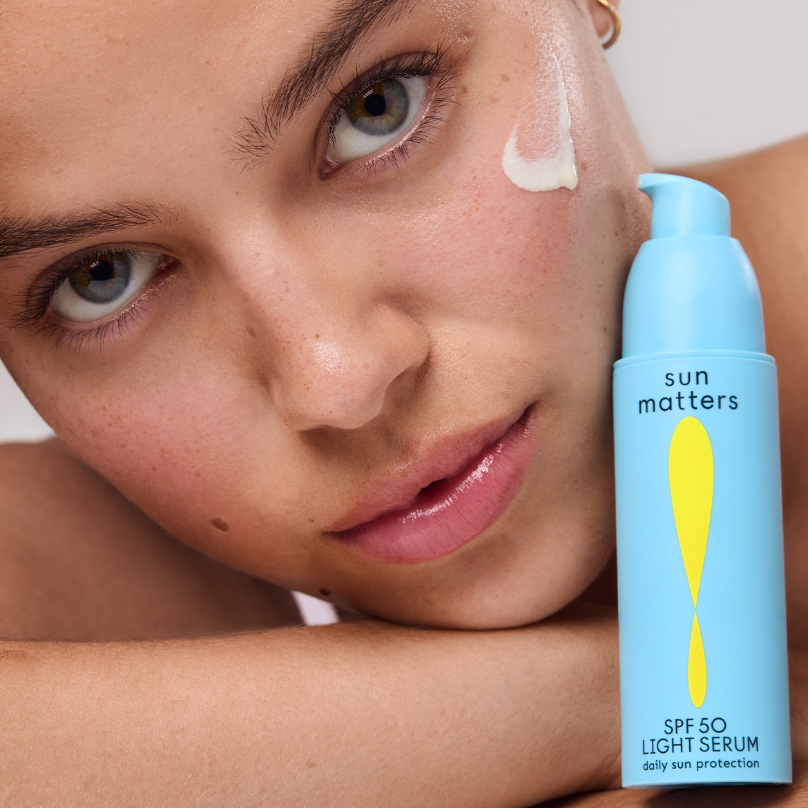 Nahaufnahme einer Frau mit einem Tropfen Sun Matters SPF 50 Light Serum auf der Wange, die blaue Flasche mit gelbem Design betont den täglichen Sonnenschutz.