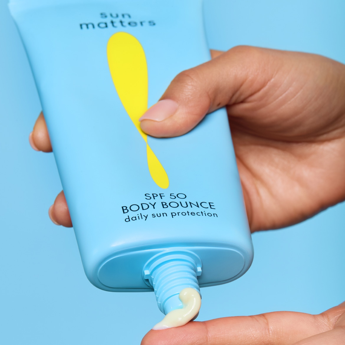 Eine Hand drückt die hellblaue „sun matters BODY BOUNCE daily sun protection SPF 50“ Sonnencreme aus der Tube auf einen Finger, wobei die cremefarbene Lotion sichtbar wird.