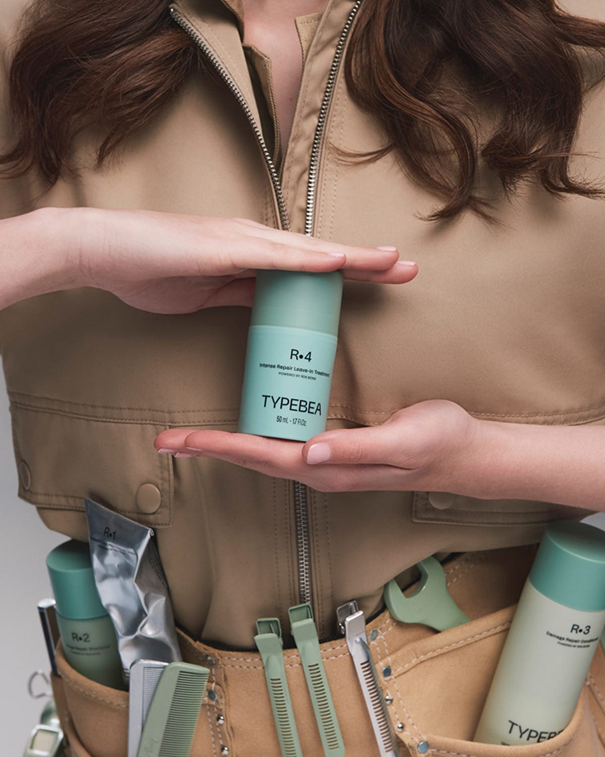 Eine Person in beigefarbener Arbeitskleidung hält eine mintgrüne Flasche TYPEBEA R.4 Intense Repair Leave-In Treatment in den Händen, während weitere Produkte und Styling-Tools in einer Werkzeugtasche sichtbar sind.