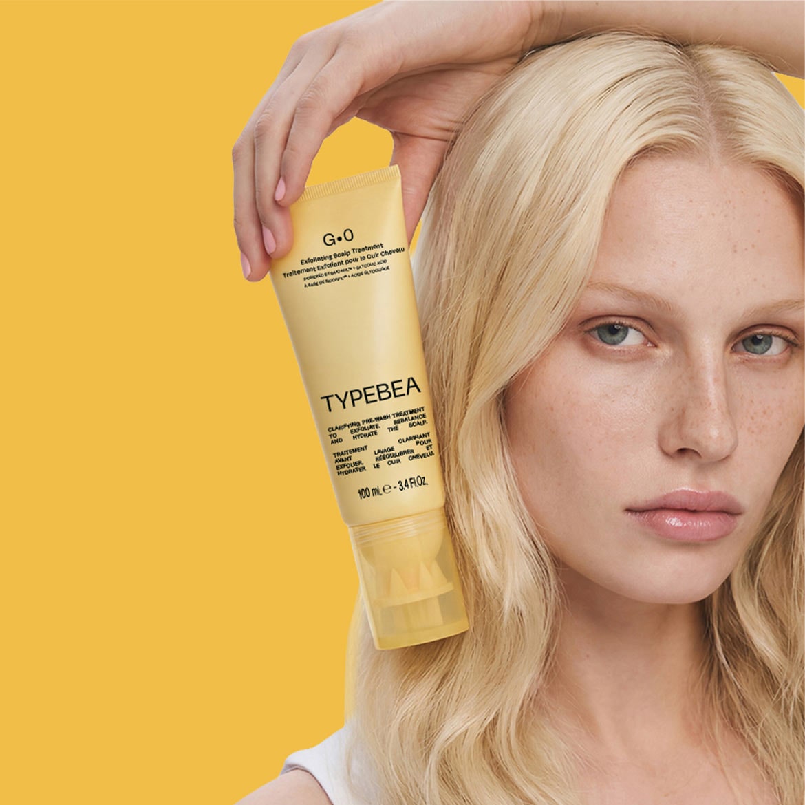 Eine Frau mit blondem Haar hält eine gelbe Tube Typebea G.0 Peeling-Kopfhautbehandlung in der Hand, die 100 ml des Produkts enthält.