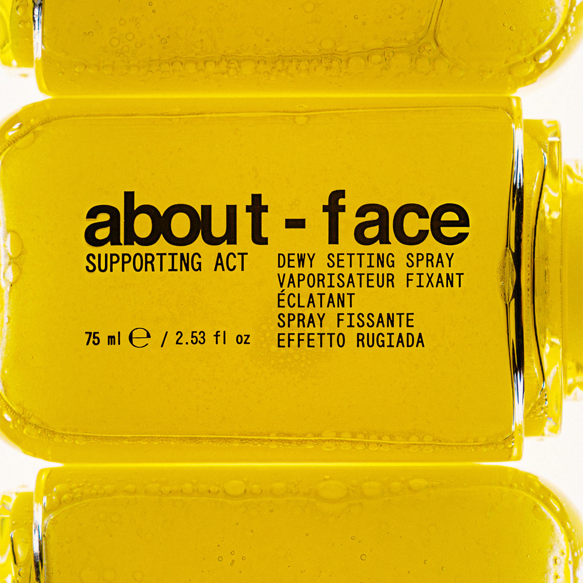 Das About-Face Supporting Act Dewy Setting Spray in einer gelben Flasche mit schwarzer Schrift fixiert Ihr Make-up und verleiht einen strahlenden Glanz.