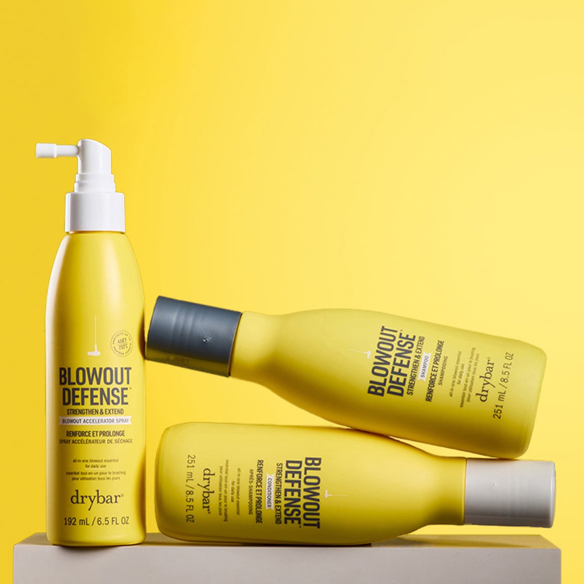 Drei gelbe Drybar Blowout Defense Produkte, darunter ein Spray mit weißem Pumpspender, ein Shampoo und ein Conditioner, liegen oder stehen auf einem hellen Untergrund vor gelbem Hintergrund.