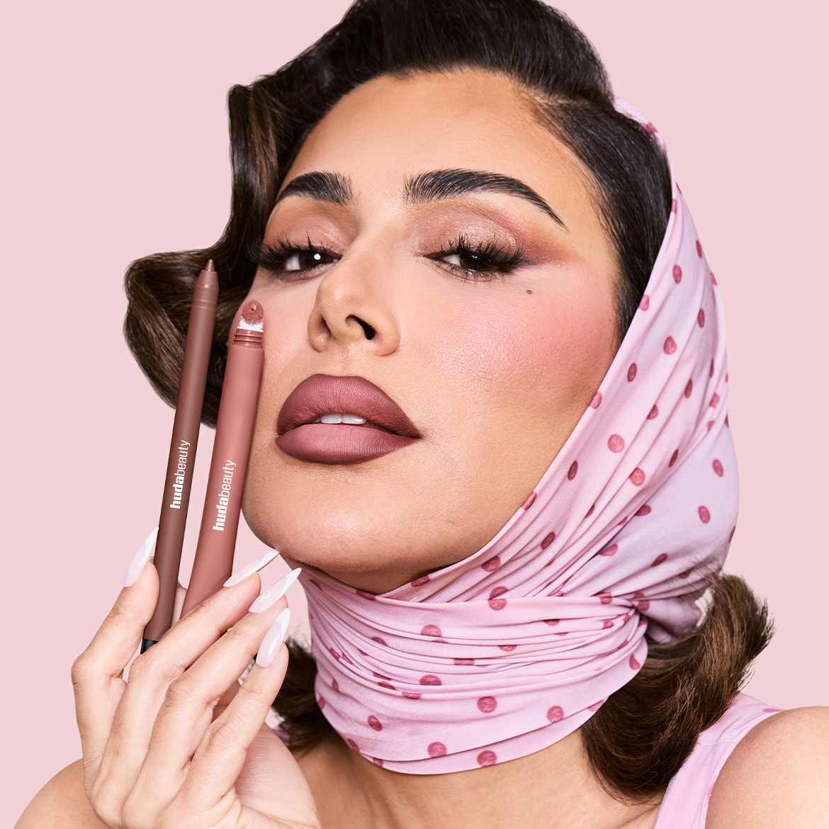 Eine Frau mit einem rosa gepunkteten Kopftuch hält zwei Huda Beauty Lippenprodukte in den Farben Braun und Nude, die ihre perfekt geschminkten Lippen ergänzen.