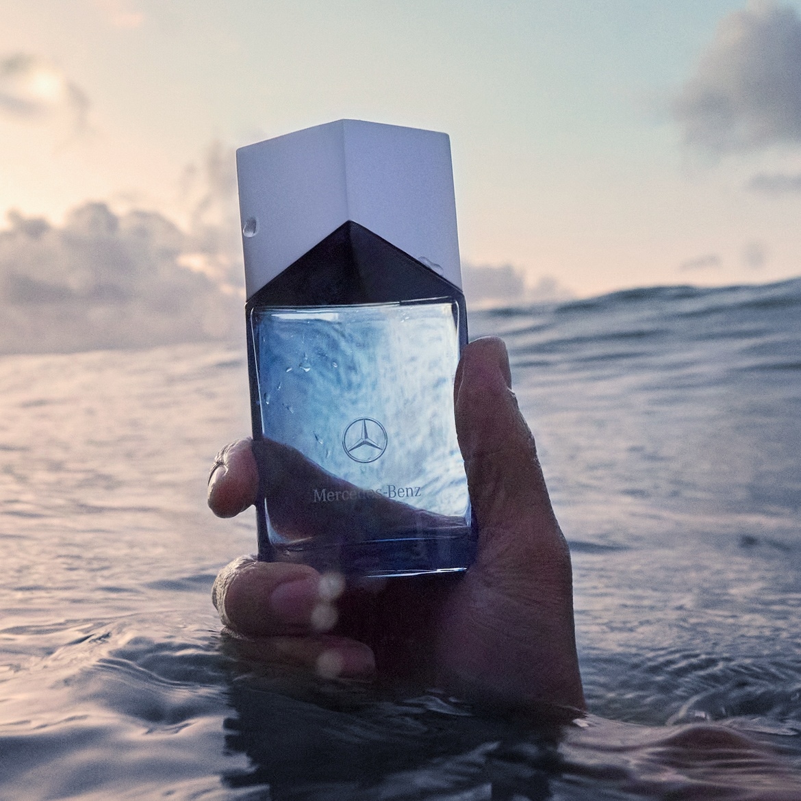 Eine Hand hält eine Flasche Mercedes-Benz-Parfüm im Wasser, der Flakon ist klar mit einem weißen Deckel, das Mercedes-Logo ist sichtbar.