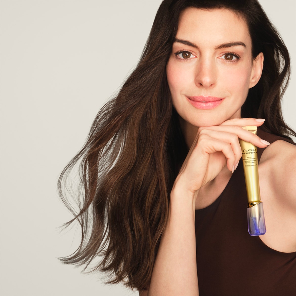 Eine Frau mit langen, gewellten, braunen Haaren hält eine goldene Shiseido-Tube mit blauem Applikator in der Hand und lächelt.