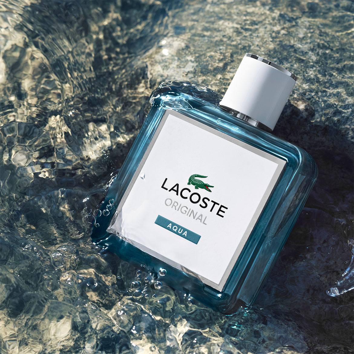 Das Lacoste Original Aqua Eau de Toilette präsentiert sich in einem durchsichtigen, hellblauen Flakon mit weißem Etikett, das den Lacoste-Schriftzug und das Krokodil-Logo zeigt, während es im Wasser liegt.
