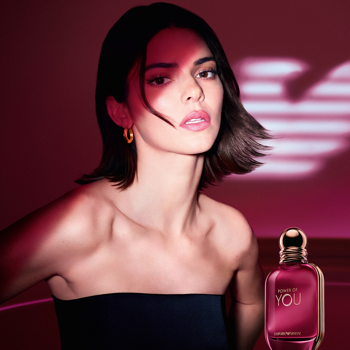 Das Bild zeigt eine Frau mit kurzem, dunklem Haar, goldenen Ohrringen und einem schwarzen trägerlosen Oberteil, neben der das Parfum 'Power of You' von Emporio Armani steht.