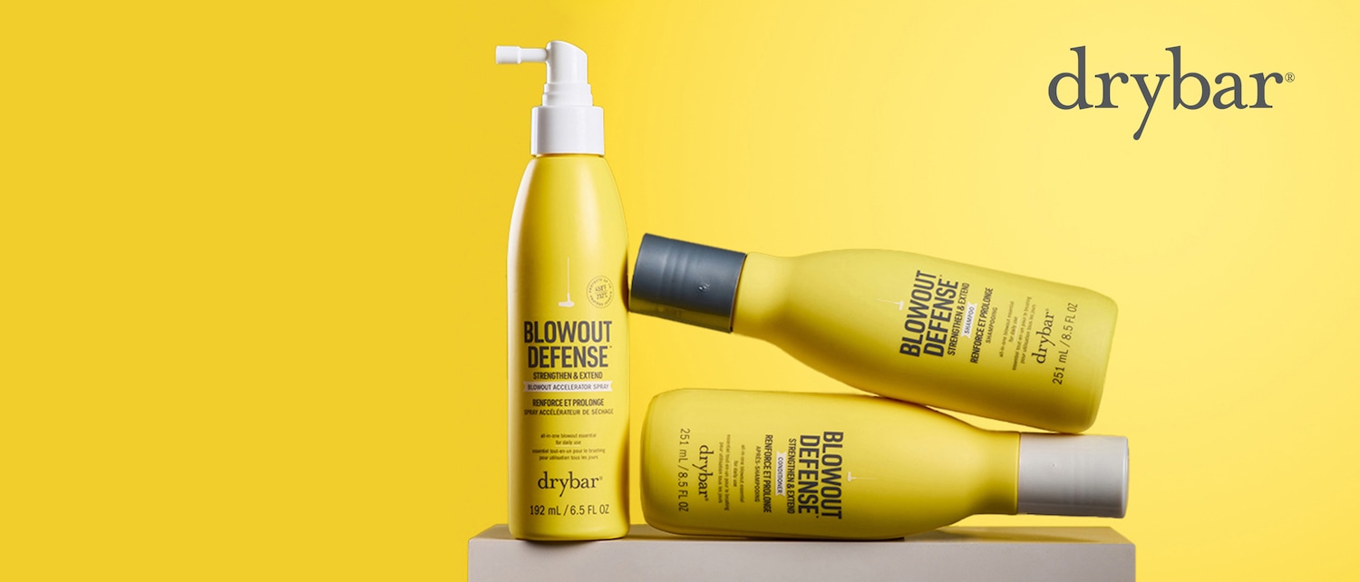 Drei gelbe Drybar Blowout Defense Produkte, darunter ein Spray, Shampoo und Conditioner, sind auf einem gelben Hintergrund angeordnet, wobei das Spray aufrecht steht und die anderen beiden schräg dahinter liegen.