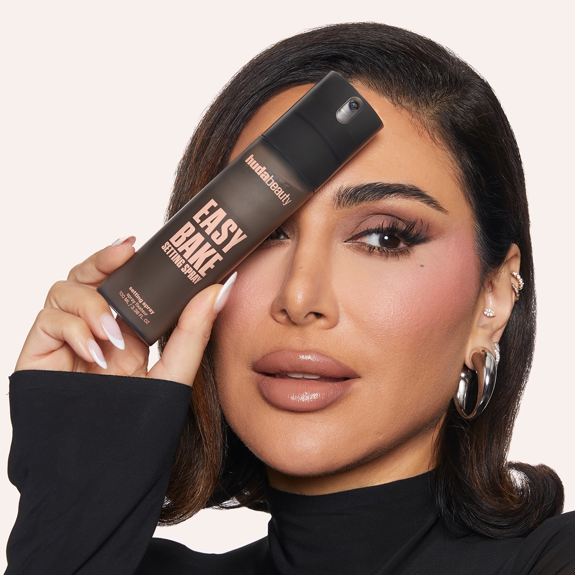 Eine Frau mit vollständigem Make-up hält eine braune Sprühflasche mit der Aufschrift „Huda Beauty Easy Bake Setting Spray“ vor ihr Gesicht.