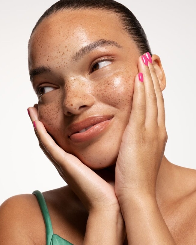 Nahaufnahme einer Frau mit Sommersprossen, die ihre Wangen berührt und einen natürlichen, strahlenden Teint zeigt, der den Trend des Skinimalismus verkörpert.