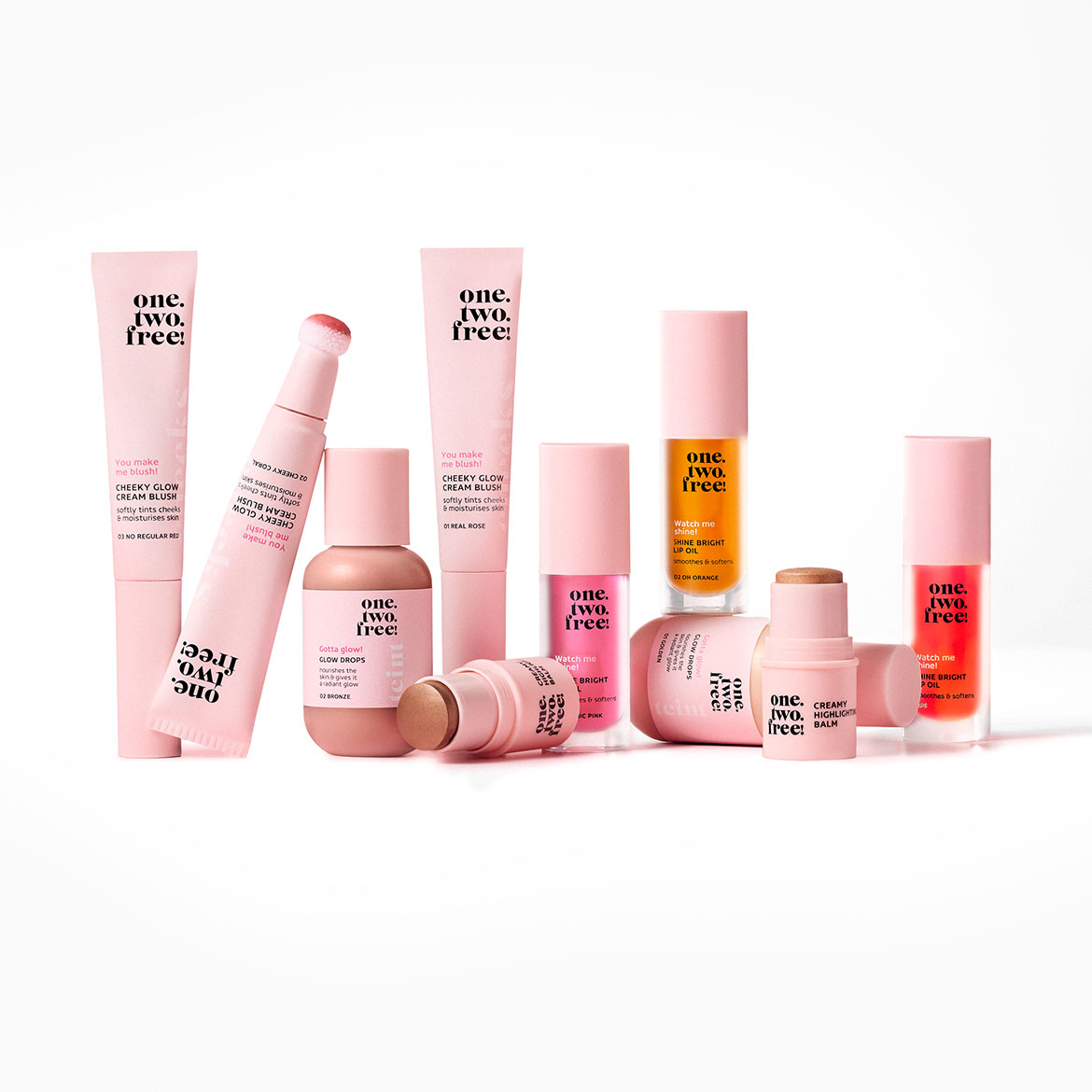 Collection of One. Two. Free! makeup products in pink packaging, including Cheeky Glow Cream Blush, Glow Drops, Shine Bright Lip Oil, and Creamy Highlighter Balm.