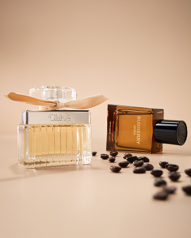 Zwei Parfümflaschen, eine von Chloé mit einer Schleife und eine von Burberry Hero, liegen auf einer beigen Oberfläche, umgeben von Kaffeebohnen.