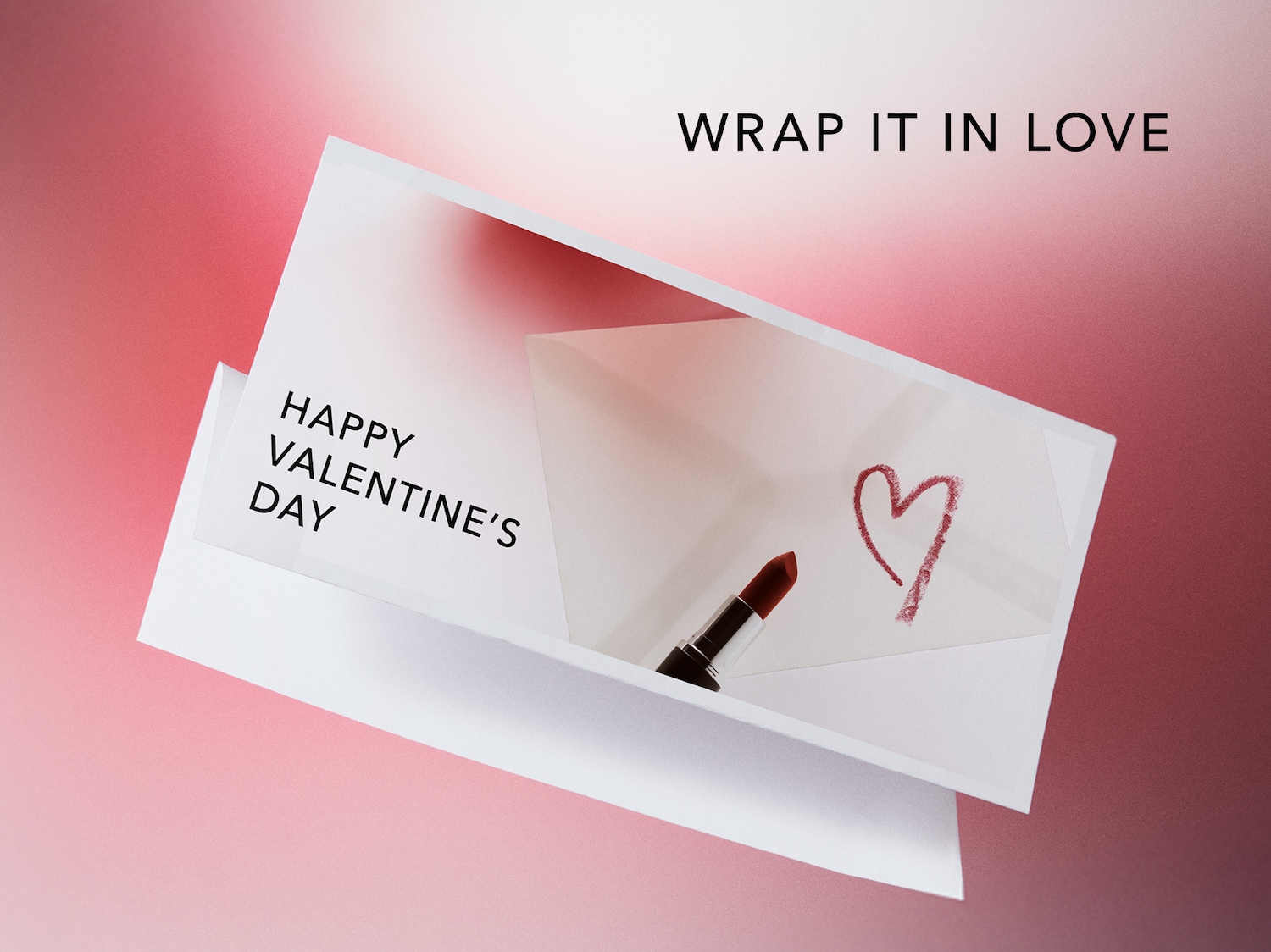 Auf einer Karte steht 'Happy Valentine's Day' neben einem roten Lippenstift und einem gezeichneten Herz, darüber der Schriftzug 'Wrap it in love'.