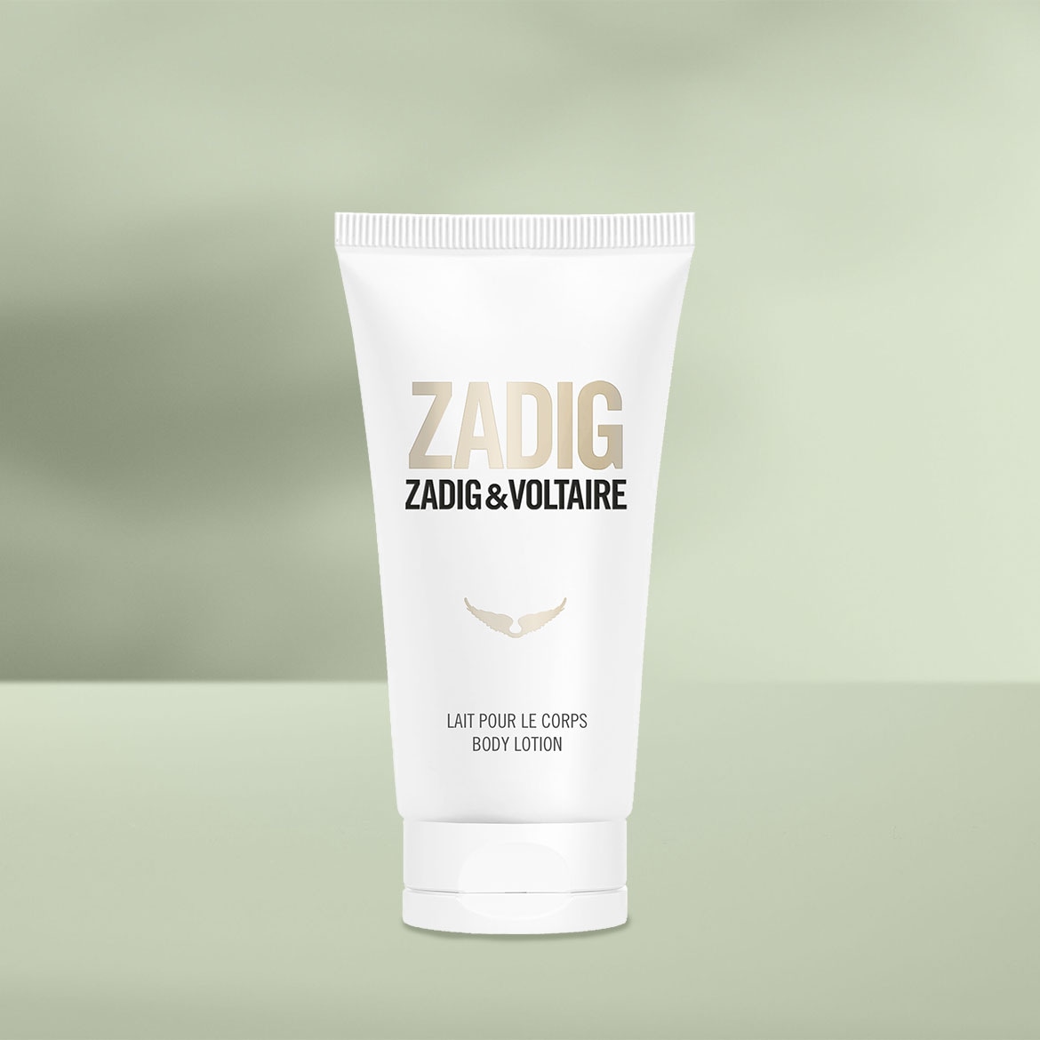 Weiße Tube Zadig&Voltaire Körperlotion mit goldener Schrift und geflügeltem Logo vor einem sanftgrünen Hintergrund.
