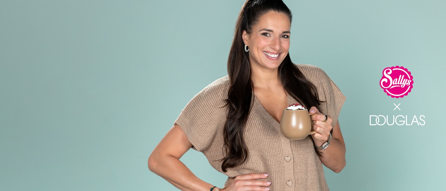 Eine lächelnde Frau mit langen, dunklen Haaren, die zu einem hohen Pferdeschwanz gebunden sind, trägt einen beigen Strickpullover und hält eine beige Tasse mit einem Getränk, das mit Sahne und roten Beeren garniert ist.