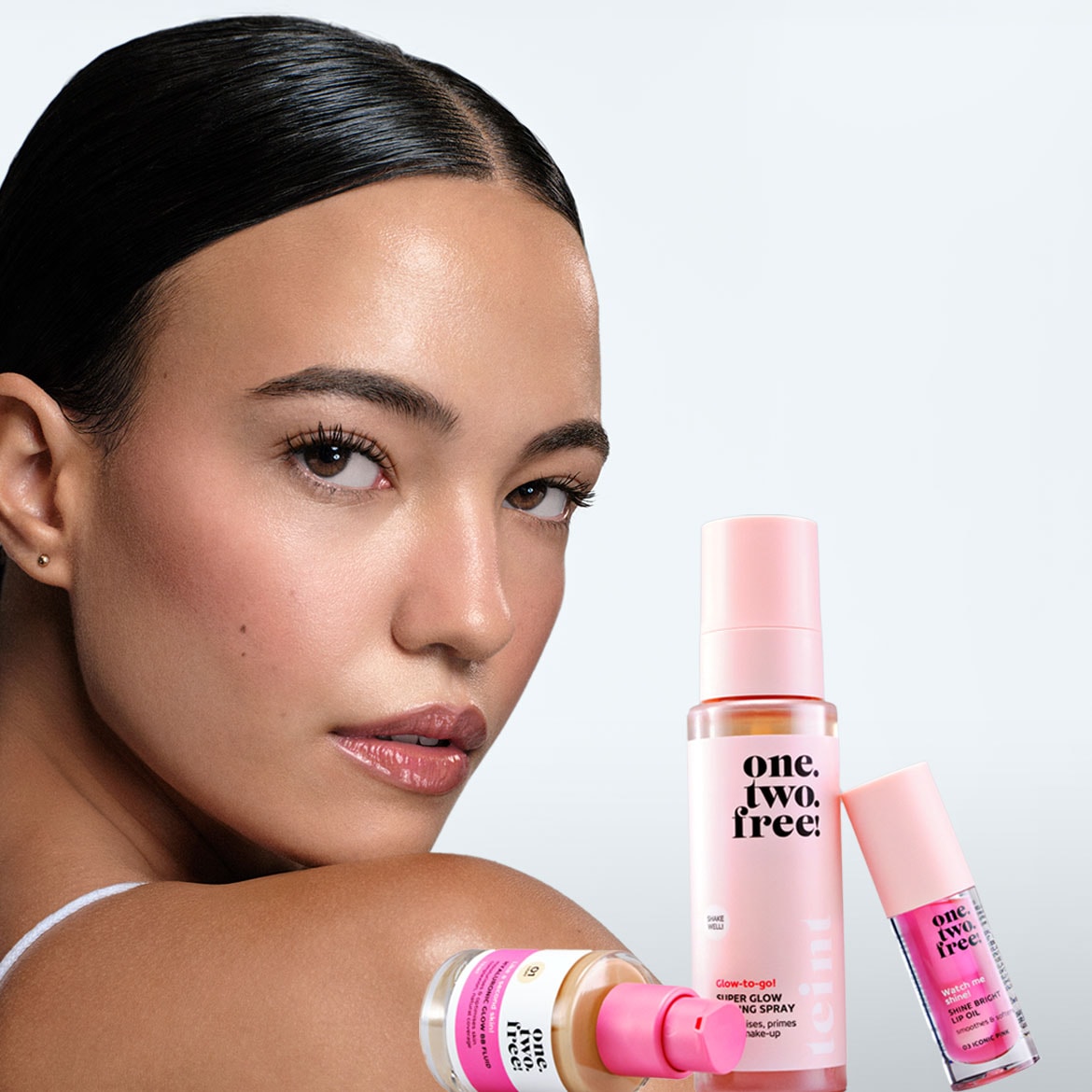 A radiant model showcases One. Two. Free! makeup: Hyaluronic Glow Fluid, Cider Glow Setting Spray, and Shine Bright Lip Oil in Iconic Pink, promising a smooth, luminous finish.