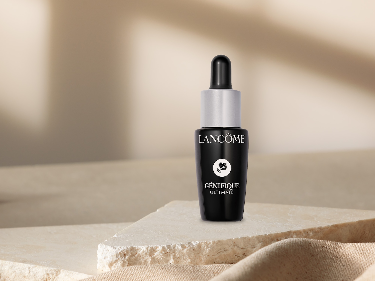 Nahaufnahme einer schwarzen Lancôme Génifique Ultimate Flasche mit silbernem Verschluss und Pipette, die auf einer hellen Steinplatte steht.