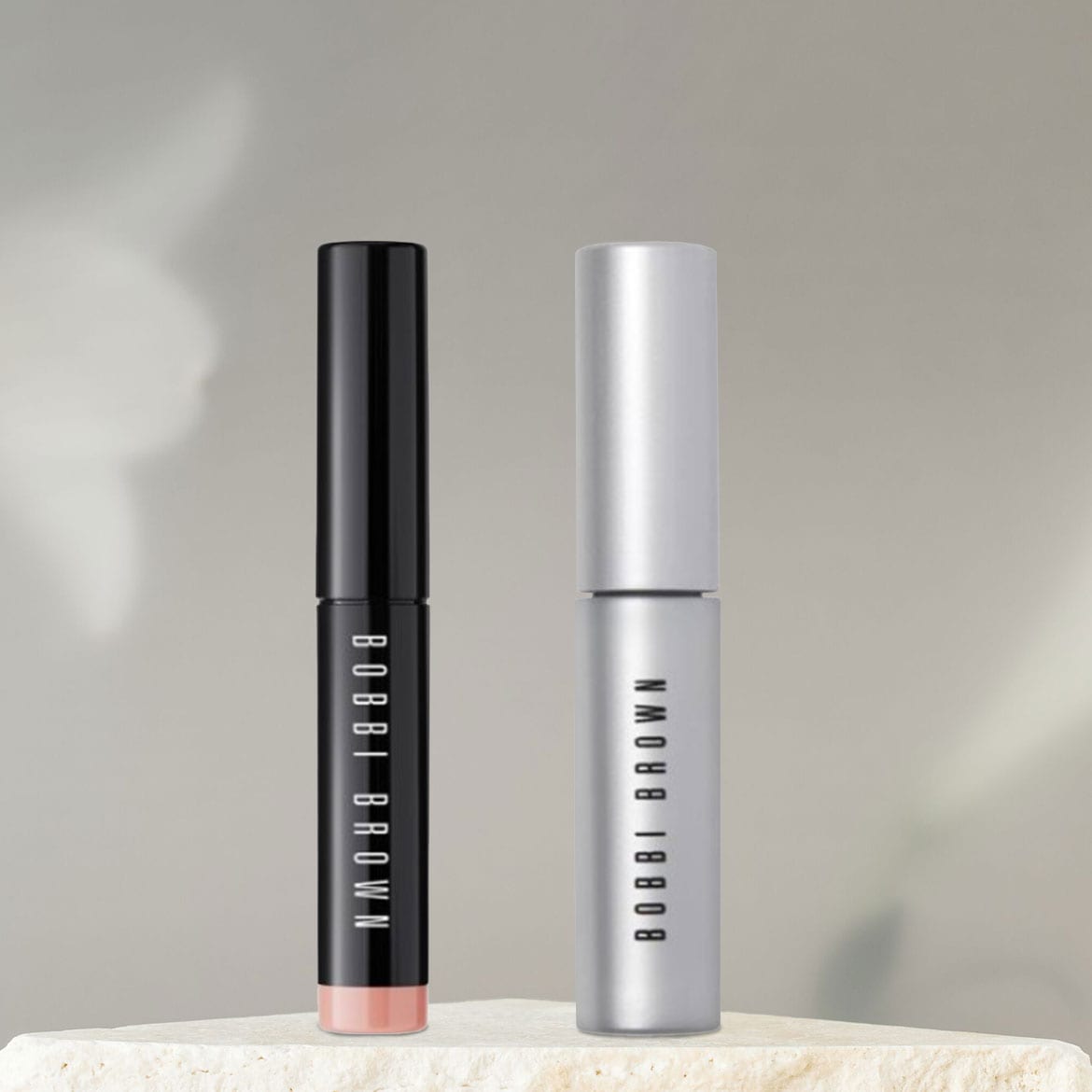 Hier sind zwei Bobbi Brown Lippenstifte, einer in einer glänzend schwarzen Hülle und der andere in einer silbernen Hülle, die auf einem cremefarbenen Sockel stehen.
