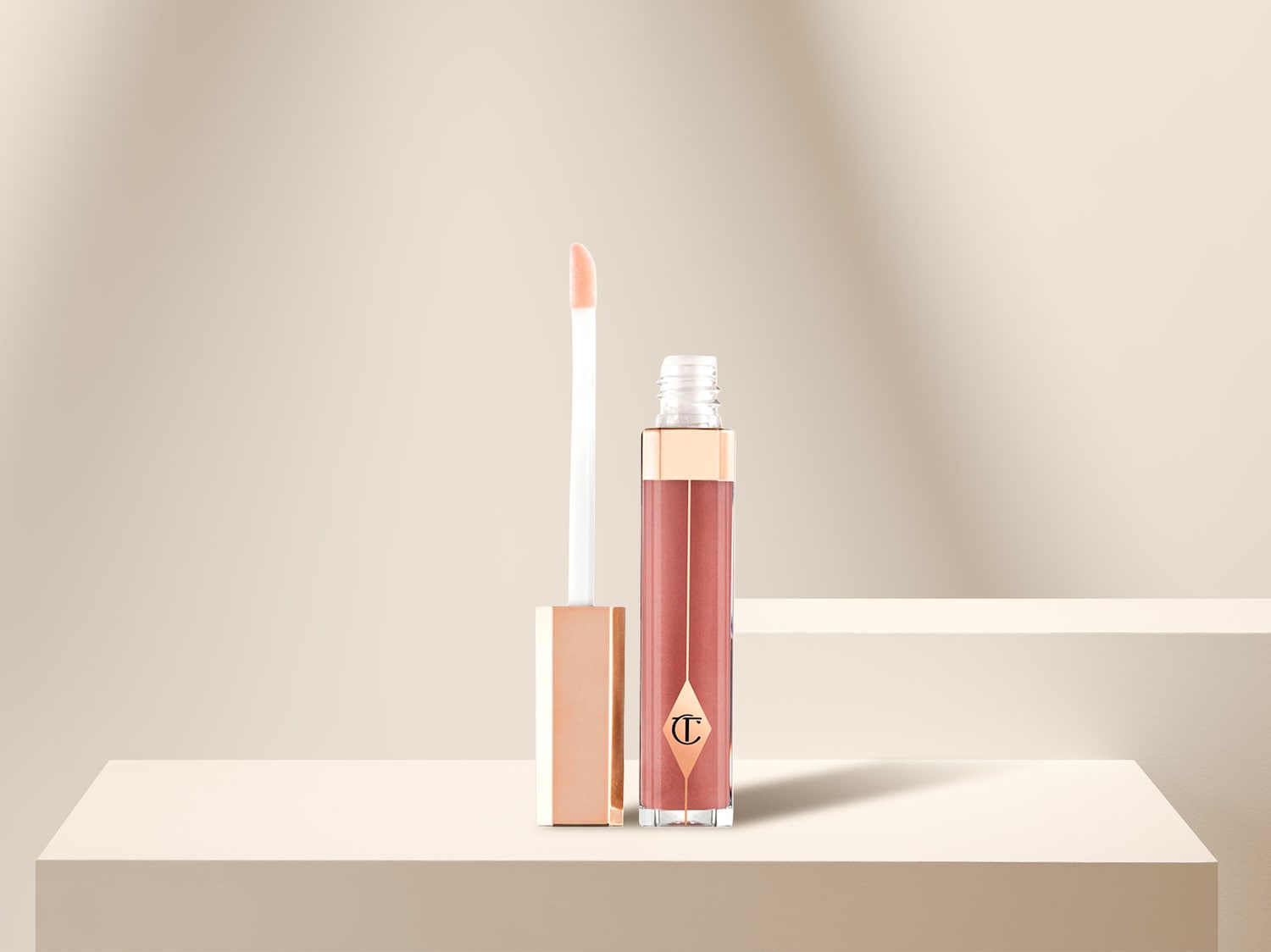 Hier ist ein glänzender Lipgloss von Charlotte Tilbury mit einem goldenen Deckel und einem Applikator, der auf einem hellen Sockel steht.