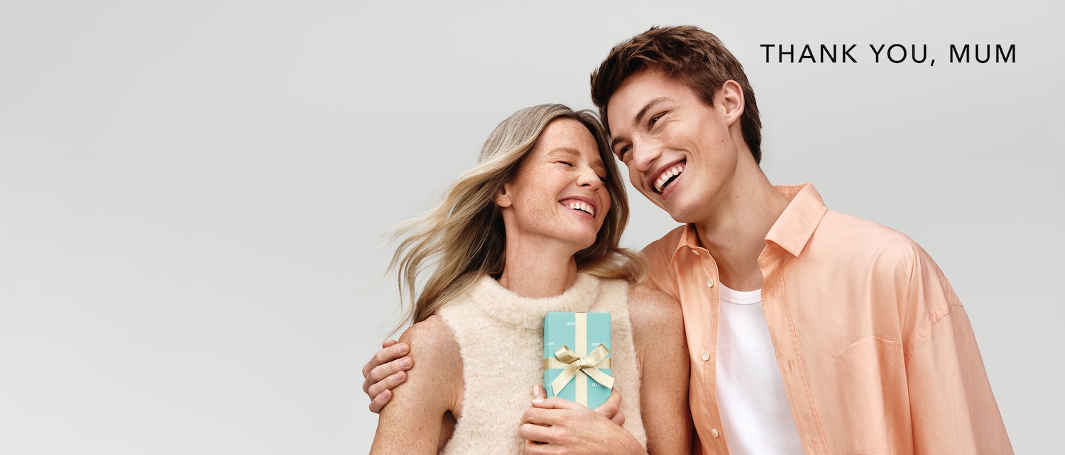 Ein strahlender Moment: Eine Mutter mit blonden Haaren und ihr Sohn umarmen sich herzlich, während sie eine hellblaue Geschenkbox mit goldener Schleife hält; darüber steht "DANKE, MAMA".