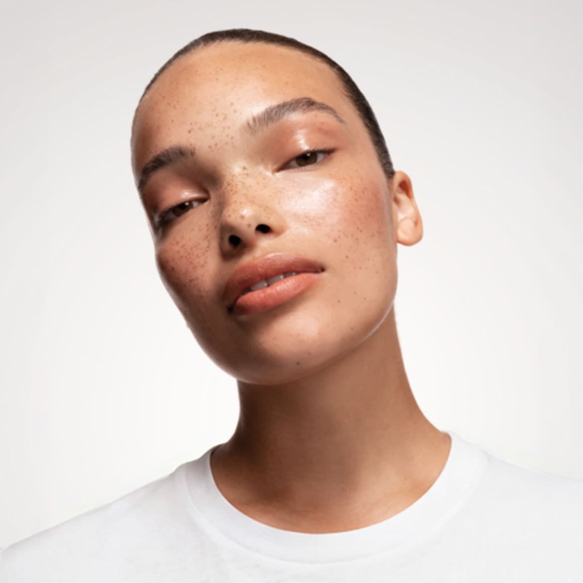 Eine junge Frau mit Sommersprossen und glänzender Haut blickt mit leicht geöffneten Lippen in die Kamera, ihr Haar ist zurückgebunden und sie trägt ein weißes T-Shirt.