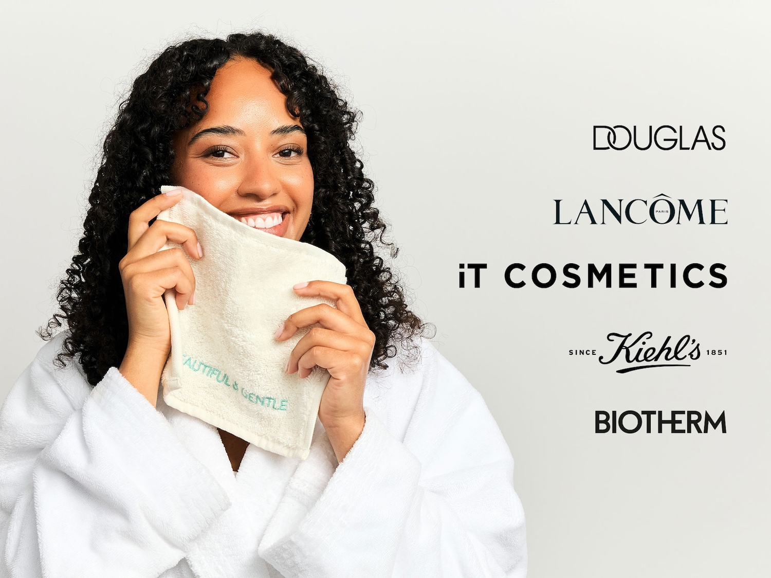 Eine lächelnde Frau mit lockigem Haar und einem weißen Bademantel hält ein cremefarbenes Handtuch mit der Aufschrift „BEAUTIFUL & GENTLE“ an ihr Gesicht.