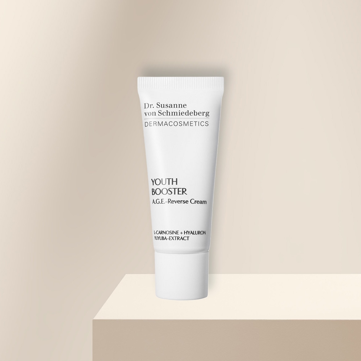 Die Dr. Susanne von Schmiedeberg YOUTH BOOSTER A.G.E.-Reverse Cream in einer weißen Tube mit schwarzer Schrift, angereichert mit L-Carnosine, Hyaluron und Yuyuba-Extrakt, steht auf einem beigefarbenen Podest.