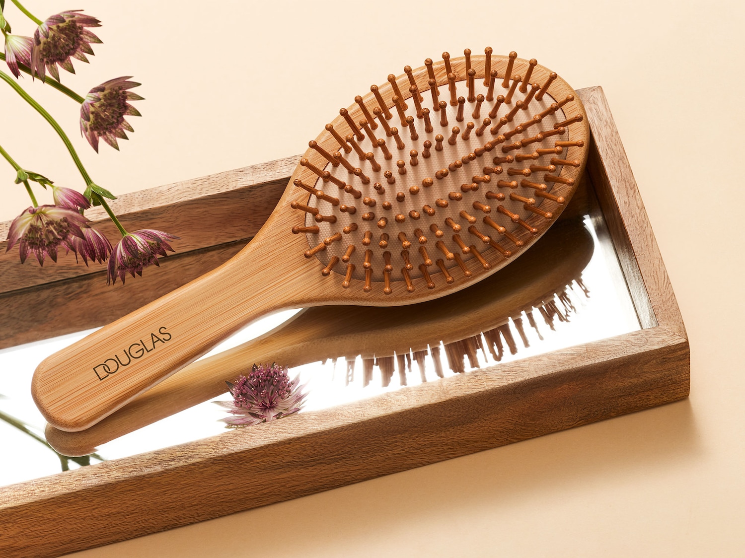 Die Douglas Haarbürste aus hellem Holz mit dem Douglas-Logo auf dem Griff liegt auf einem Spiegel in einem Holzrahmen, daneben sind lila Blumen.