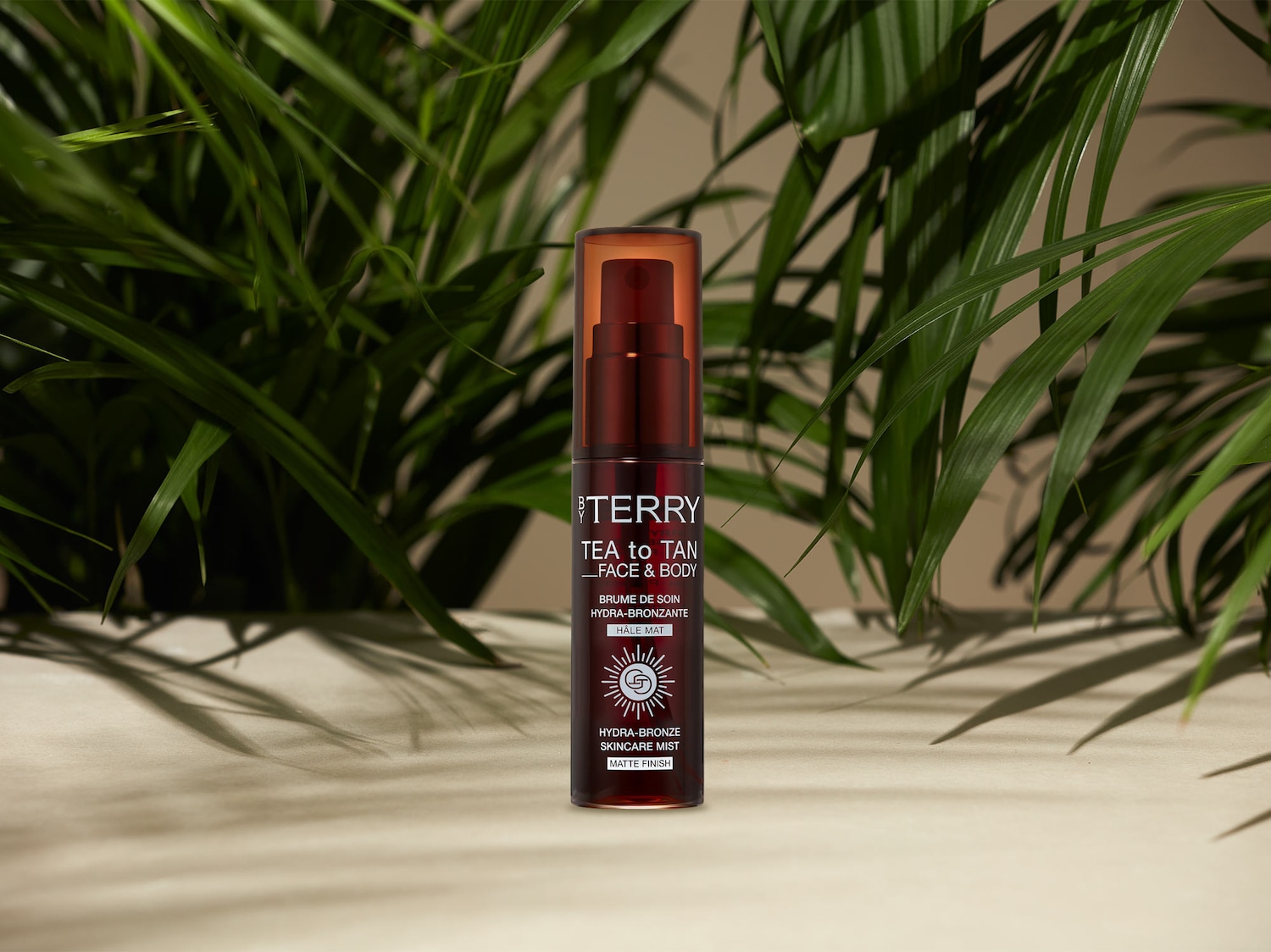 Die By Terry Tea to Tan Face & Body Hydra-Bronze Skincare Mist mit mattem Finish in einer braunen Sprühflasche steht vor einem Hintergrund aus grünen Palmblättern.
